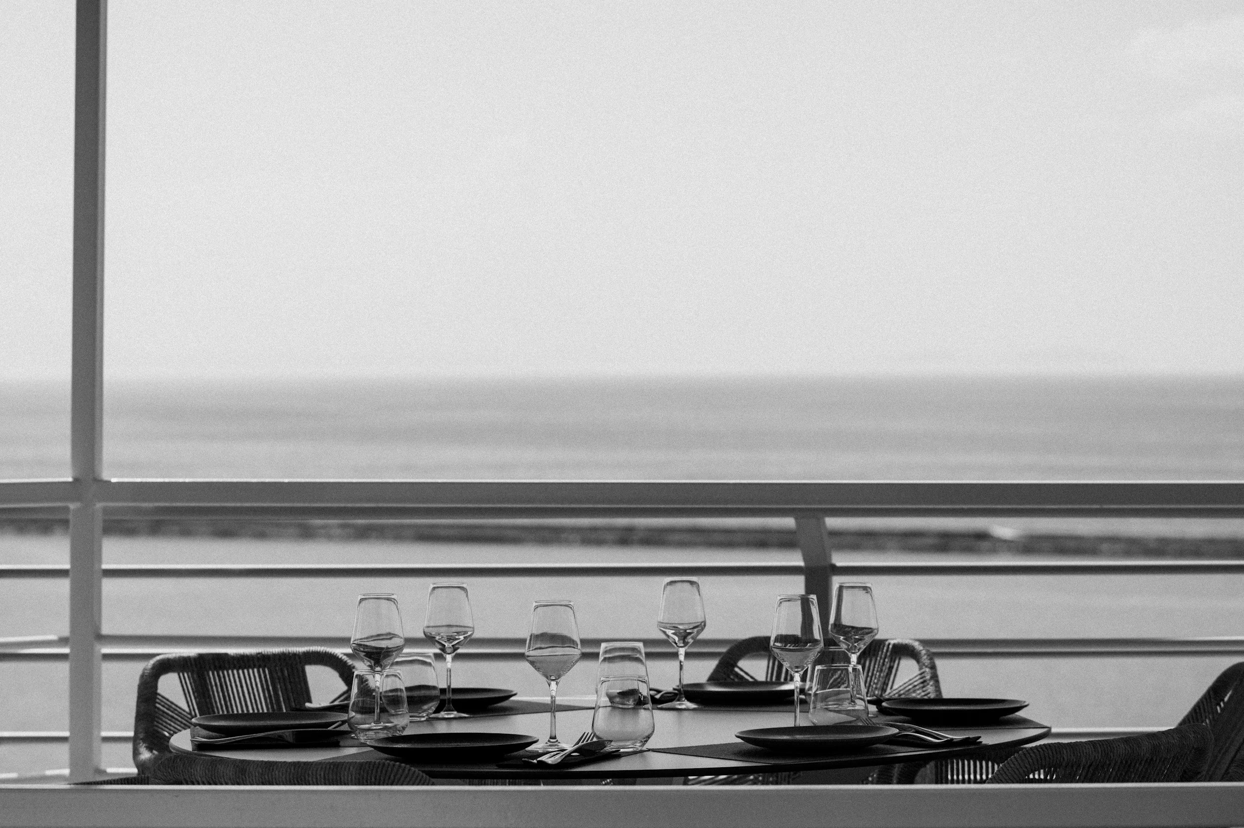 A table set for dining with wine glasses, plates, and utensils on a balcony overlooking the ocean in black and white.