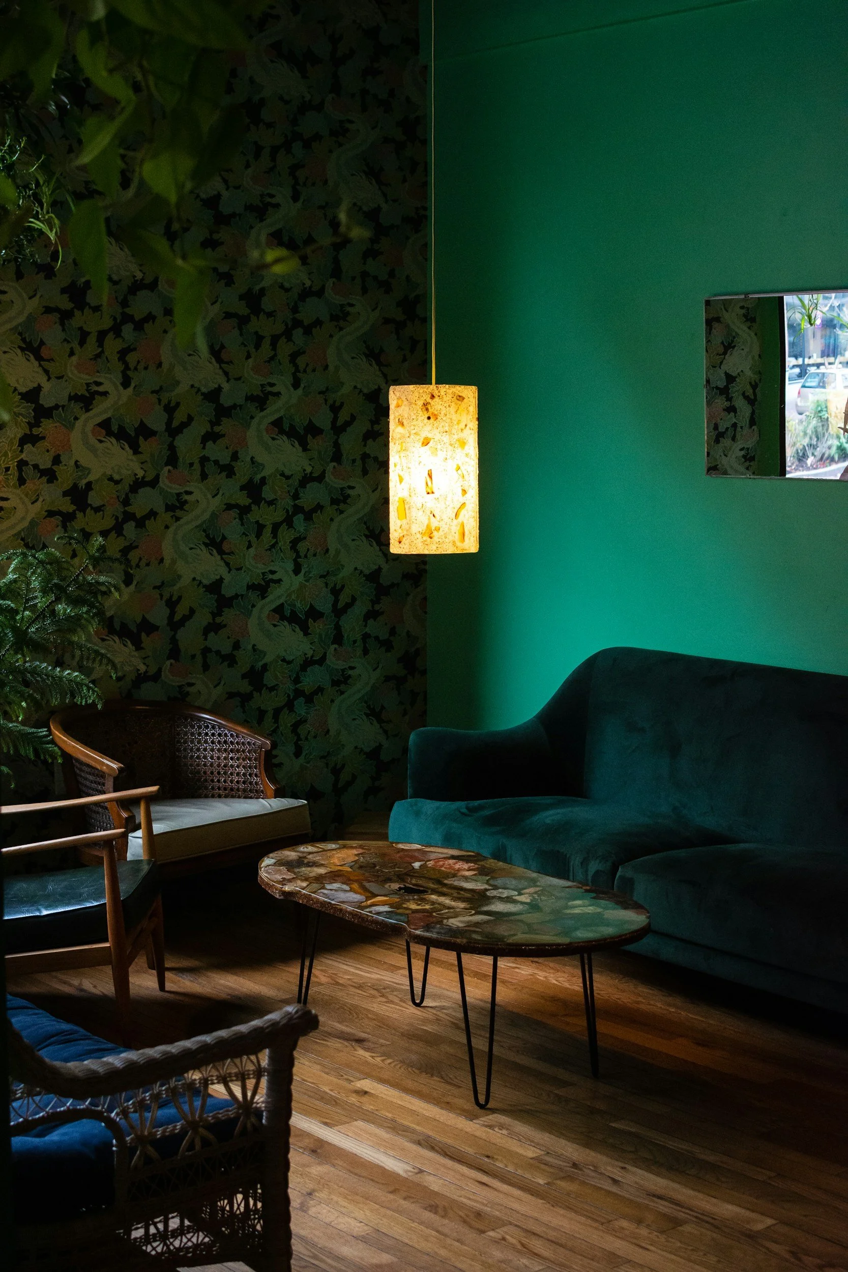 A cozy indoor space with a green velvet sofa, a wooden armchair with layered cushions, and a marble-topped coffee table with black metal legs. The walls are decorated with patterned wallpaper featuring snakes and foliage on the left, and a solid teal color on the right. A warm hanging lamp provides soft lighting, and a window on the right side shows a glimpse of outdoor cars and greenery.