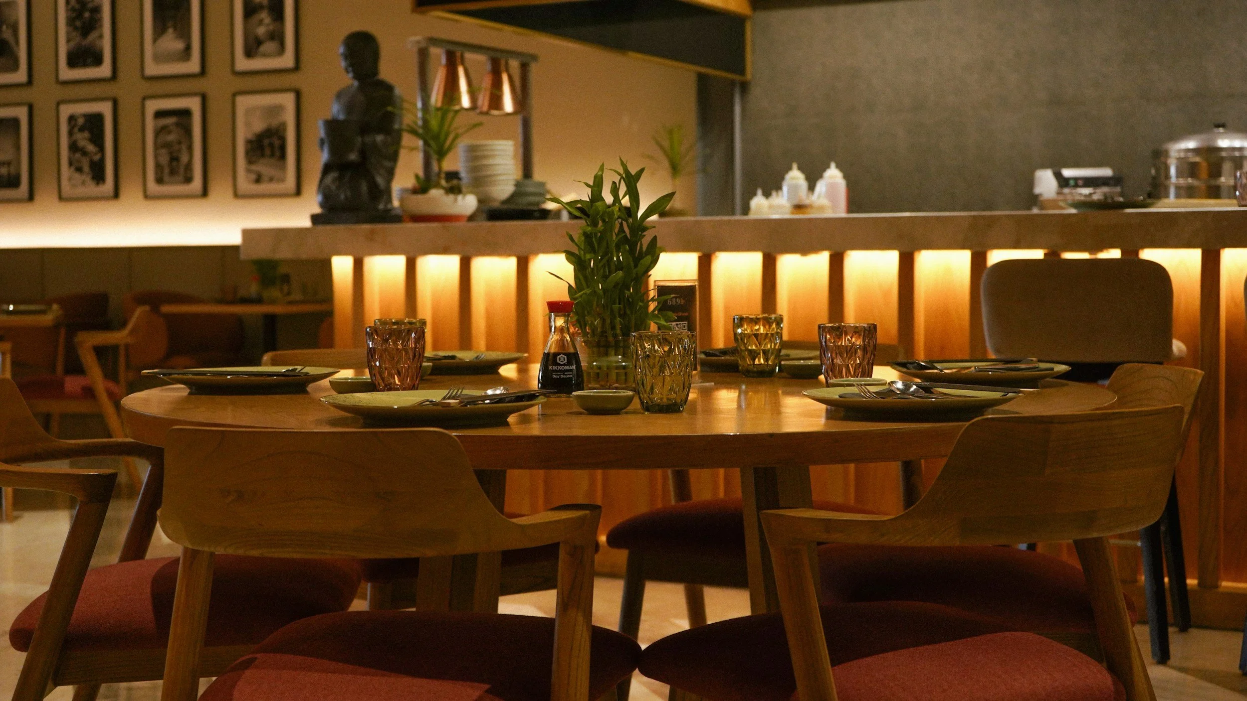 Indoor restaurant dining table set with plates, glasses, napkins, and a plant centerpiece. Behind is a counter with various condiments and decoration, with artwork on the wall.