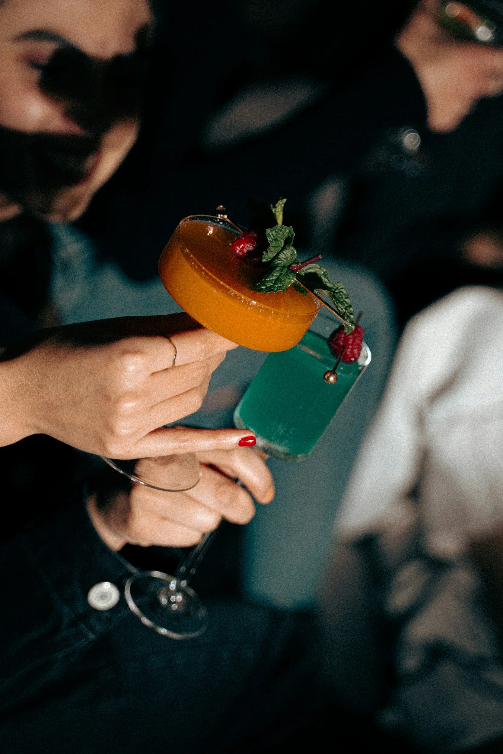 Close-up of a person holding a cocktail glass with an orange-colored drink garnished with mint leaves and berries, with a dark background.