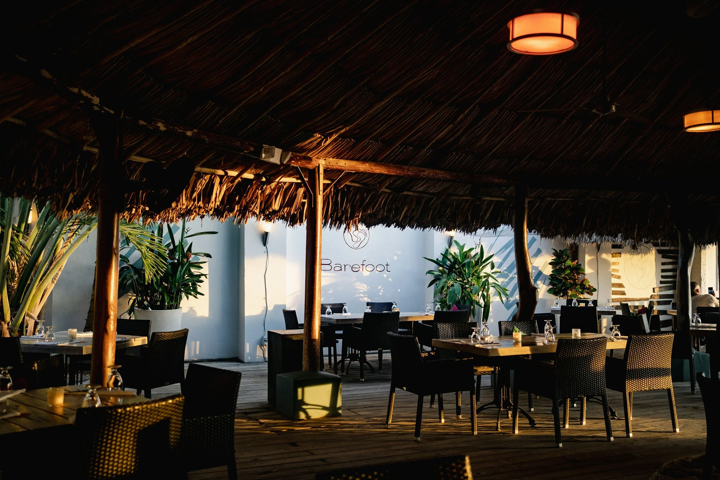 An outdoor dining area with tables and chairs under a thatched roof with warm lighting, surrounded by plants and a sign that reads "Barefoot."
