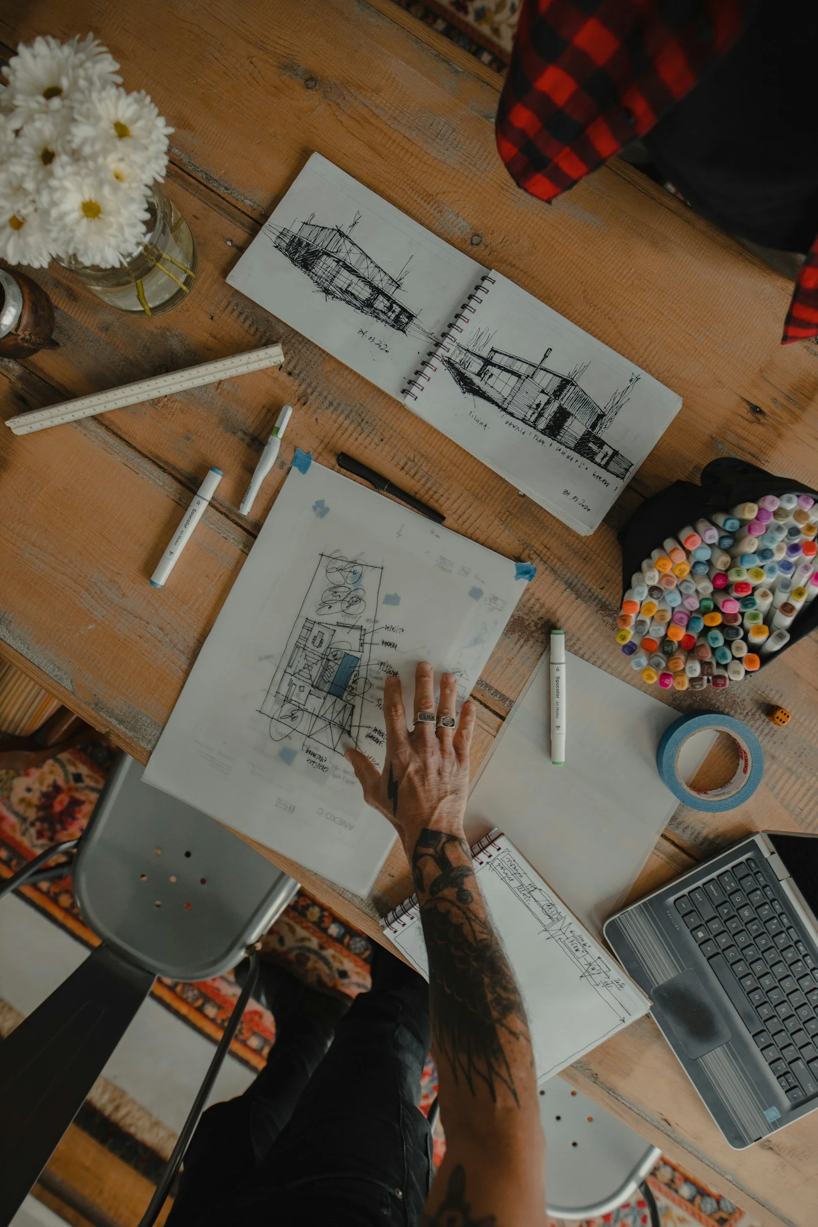 A wooden table with sketchbooks, drawings, assorted markers, pens, a laptop, tape, a ruler, a calculator, a jar of white daisies, and a person's hand with tattoos. The person is wearing black pants and a tattoo sleeve, and there is a floral rug underneath and a grey chair at the edge of the image.