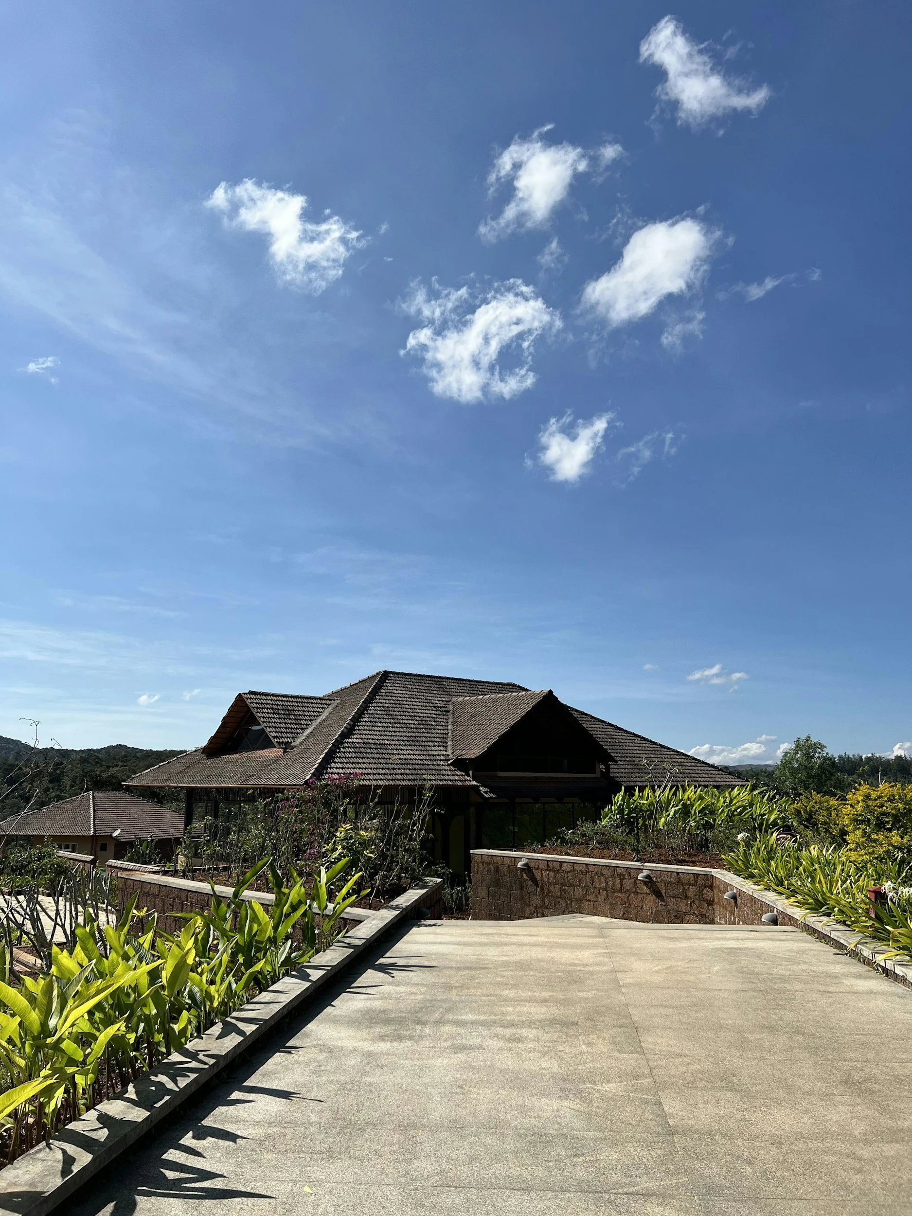 A large house with a tiled roof, surrounded by greenery, on a bright sunny day with a blue sky and some clouds.