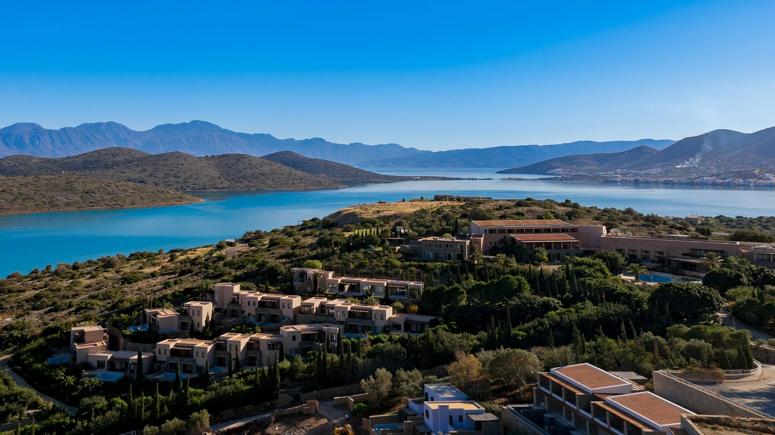 Scenic view of a lake with surrounding mountains, residential buildings, and lush greenery under a clear blue sky.
