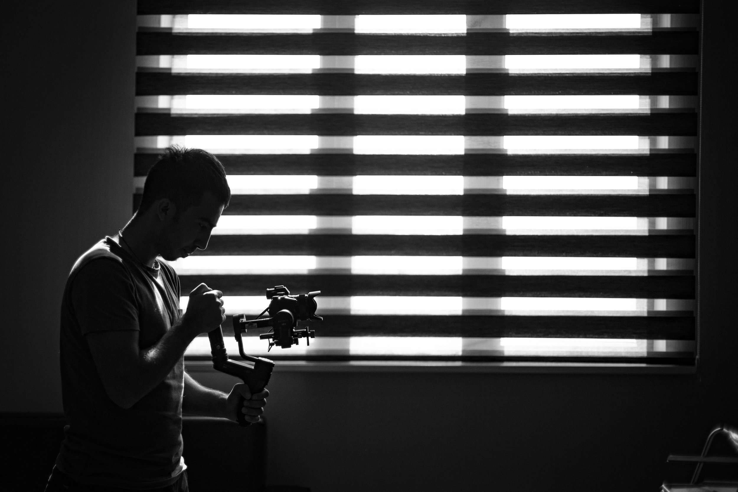 Silhouette of a person operating a camera stabilizer in a room with horizontal blinds on a window, in black and white.