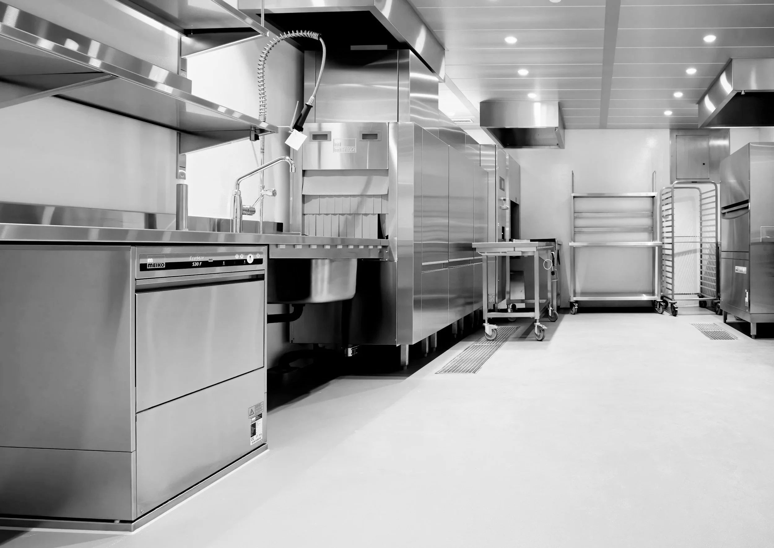 Empty commercial kitchen with stainless steel appliances, counters, and storage carts, black and white photo.