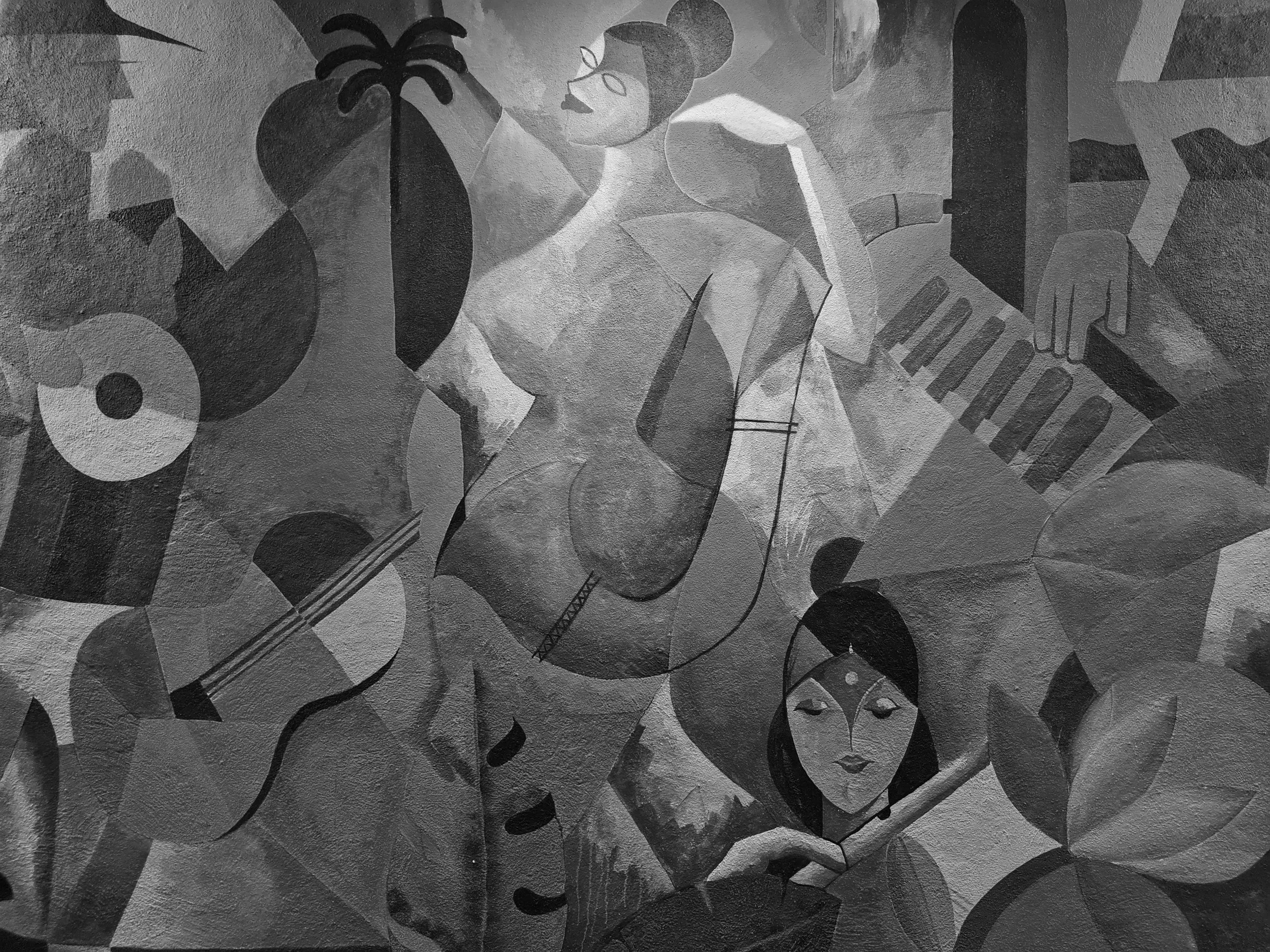 A mural of a woman with dark hair and closed eyes, holding a paintbrush, surrounded by leaves, a guitar, a brick wall, and various abstract shapes in a cubist style.