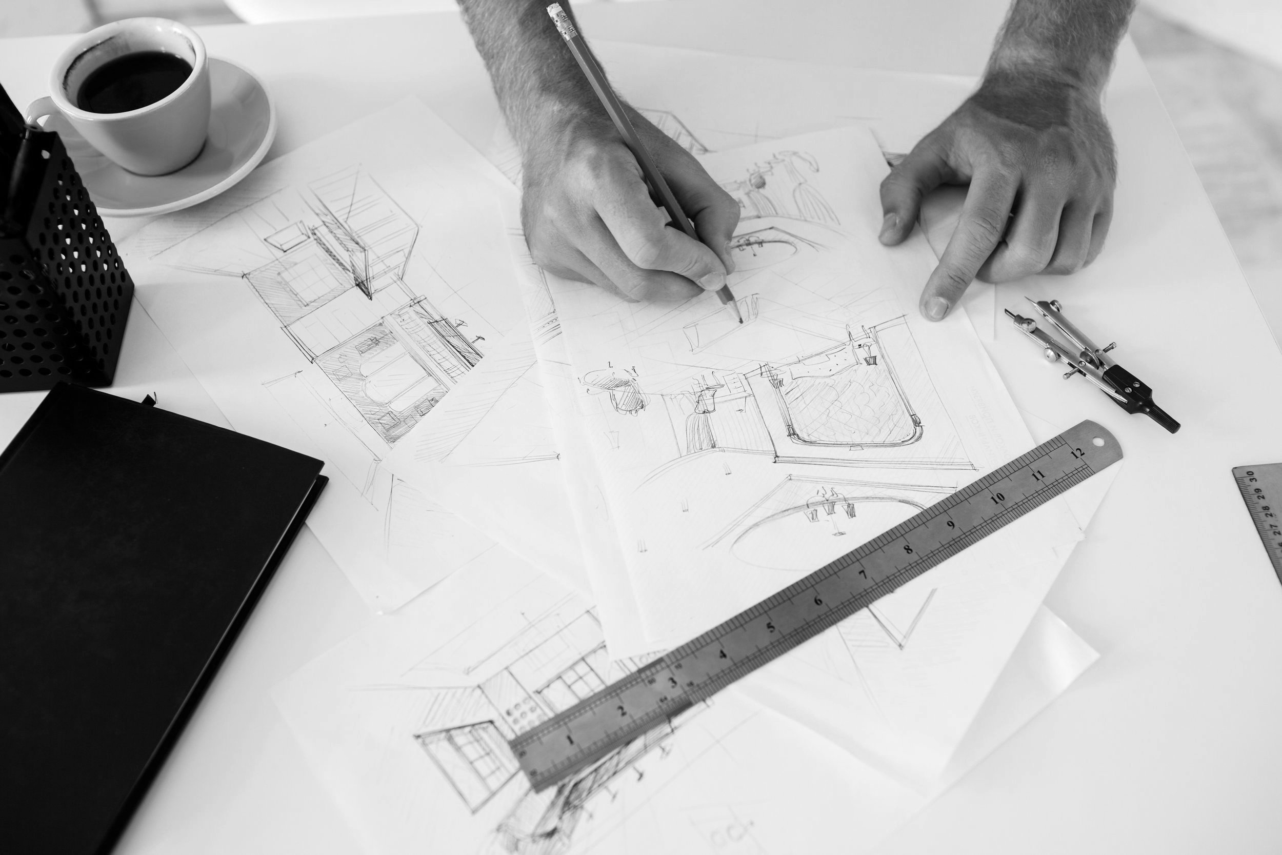 A person working on architectural sketches and blueprints at a desk with drawing tools, ruler, and a cup of coffee.