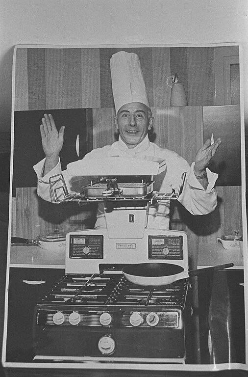 A chef in a tall white hat and apron smiling and waving in a kitchen with a stove and cooking utensils.