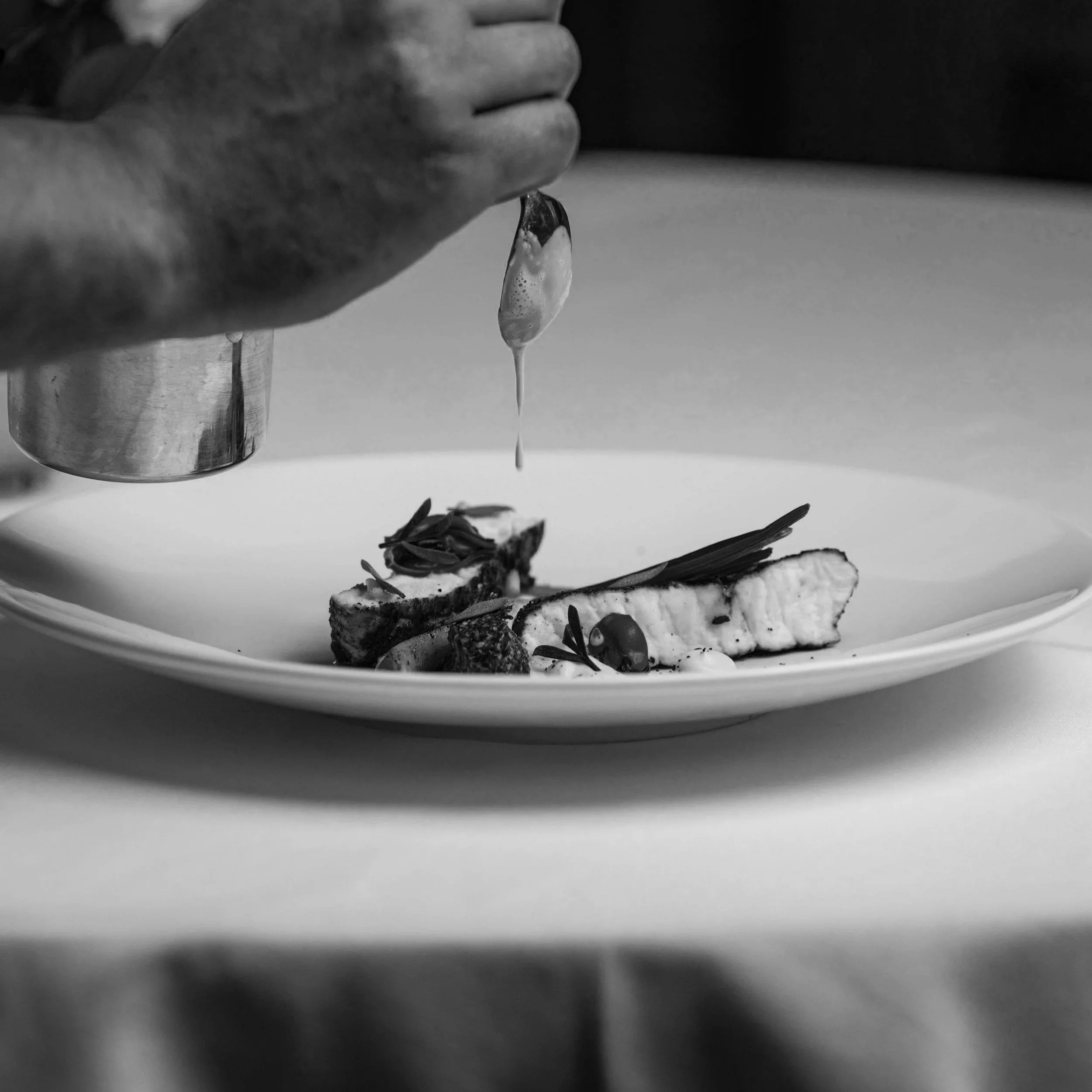 A chef is drizzling sauce or oil onto a plated gourmet dish with grilled fish, garnished with herbs and vegetables, in a professional kitchen setting.
