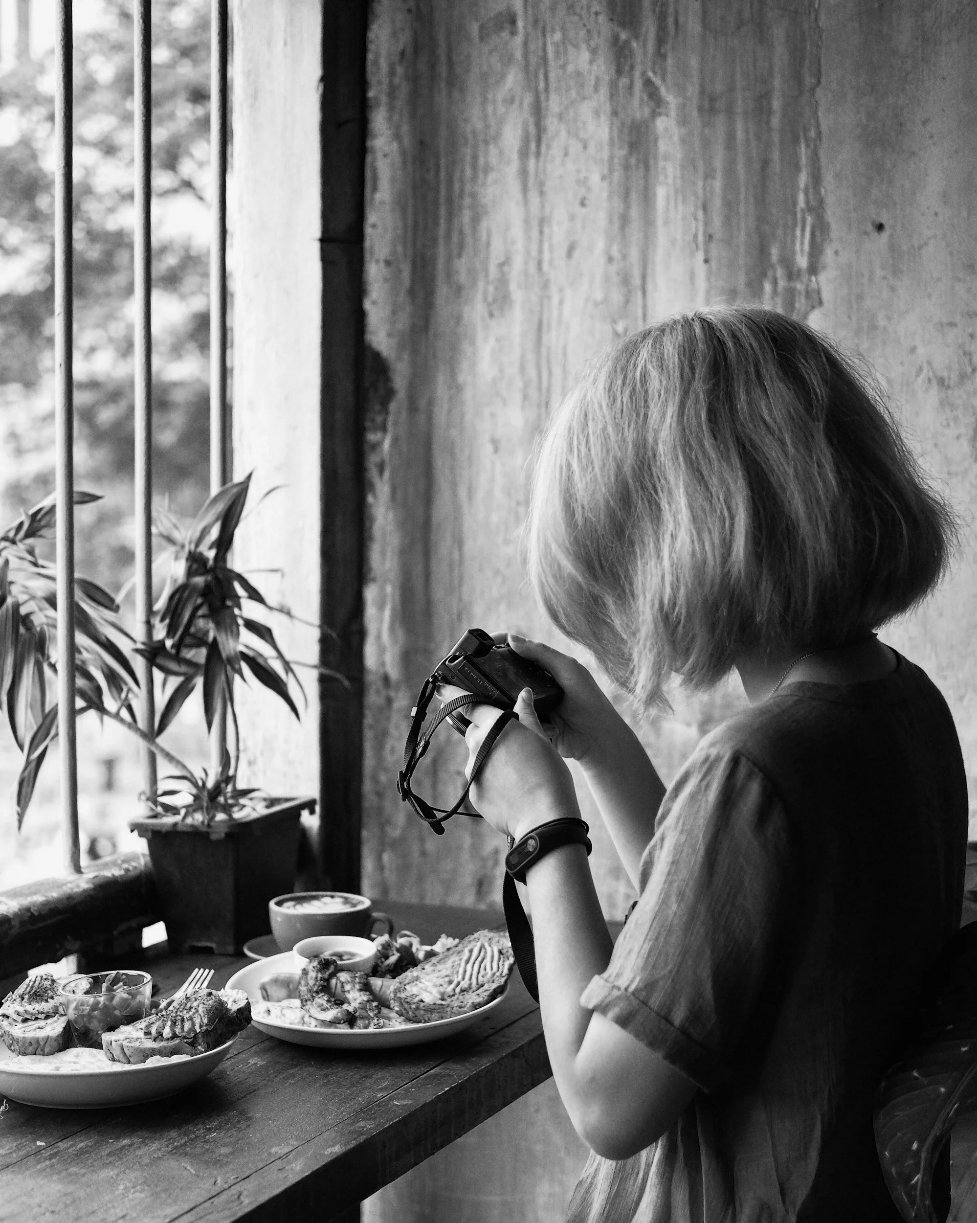 A woman with shoulder-length hair taking a photo with a camera at a table with food in a rustic setting