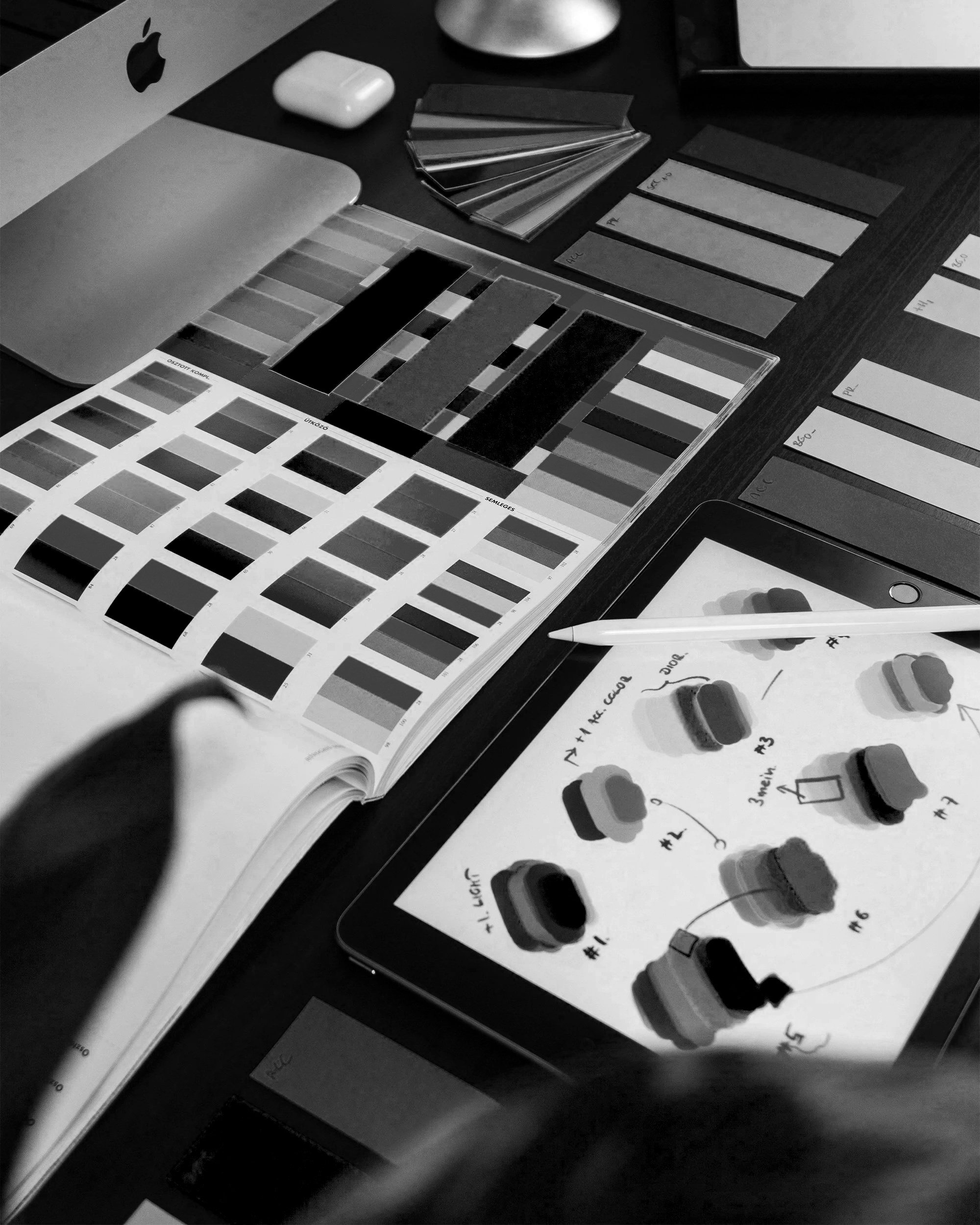 A workspace with color swatches, paint samples, and notes on a tablet, with a laptop and AirPods nearby.