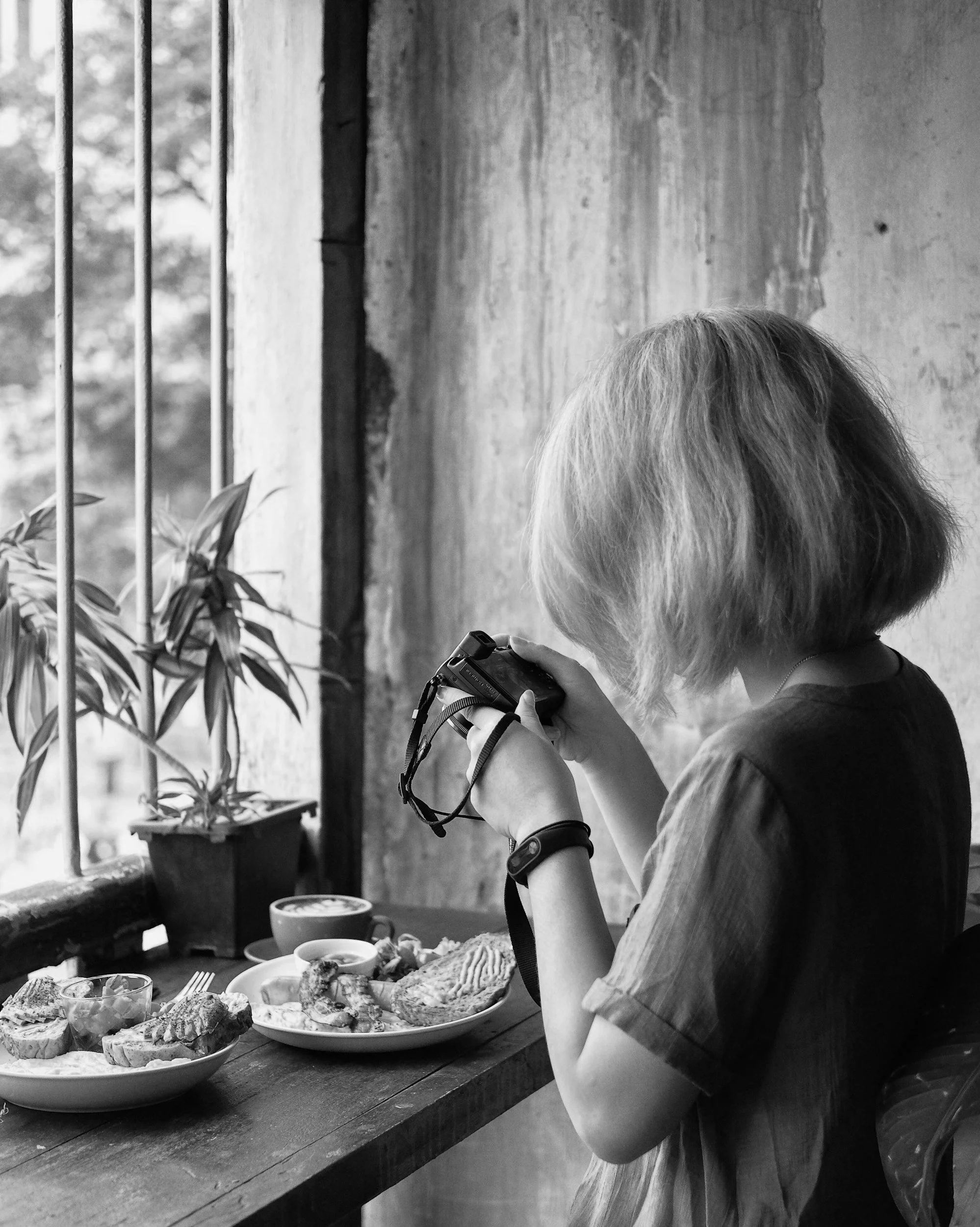 A person with light-colored hair is sitting at a wooden table in a rustic setting, eating a meal. The table has several plates of food, and the person is holding a camera, possibly taking a picture of the food. In the background, there are plants, a window with bars, and a concrete wall.