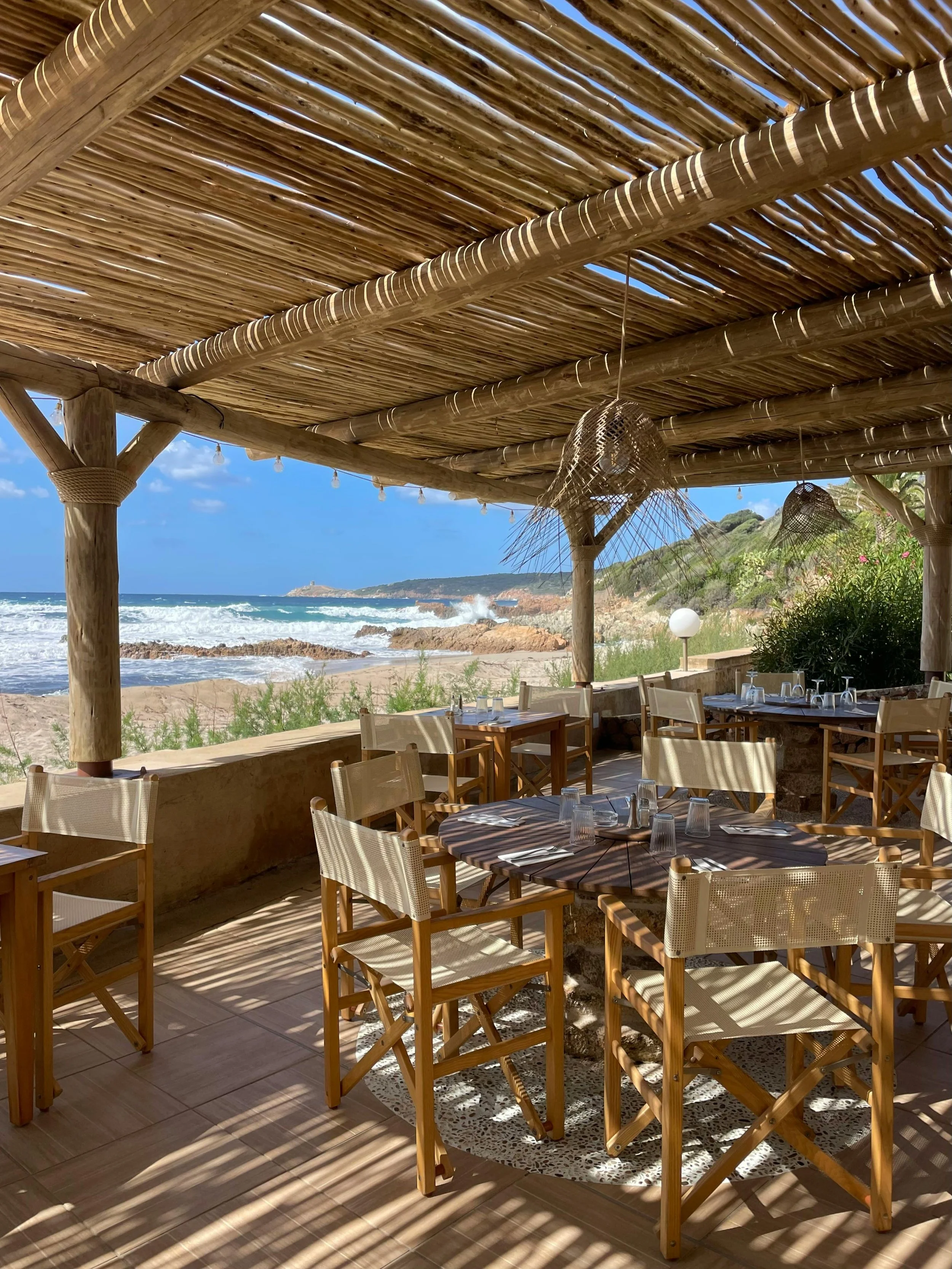 Beachside outdoor restaurant with wooden tables and chairs under a bamboo pergola, overlooking the ocean with waves and rocky shoreline in the background.