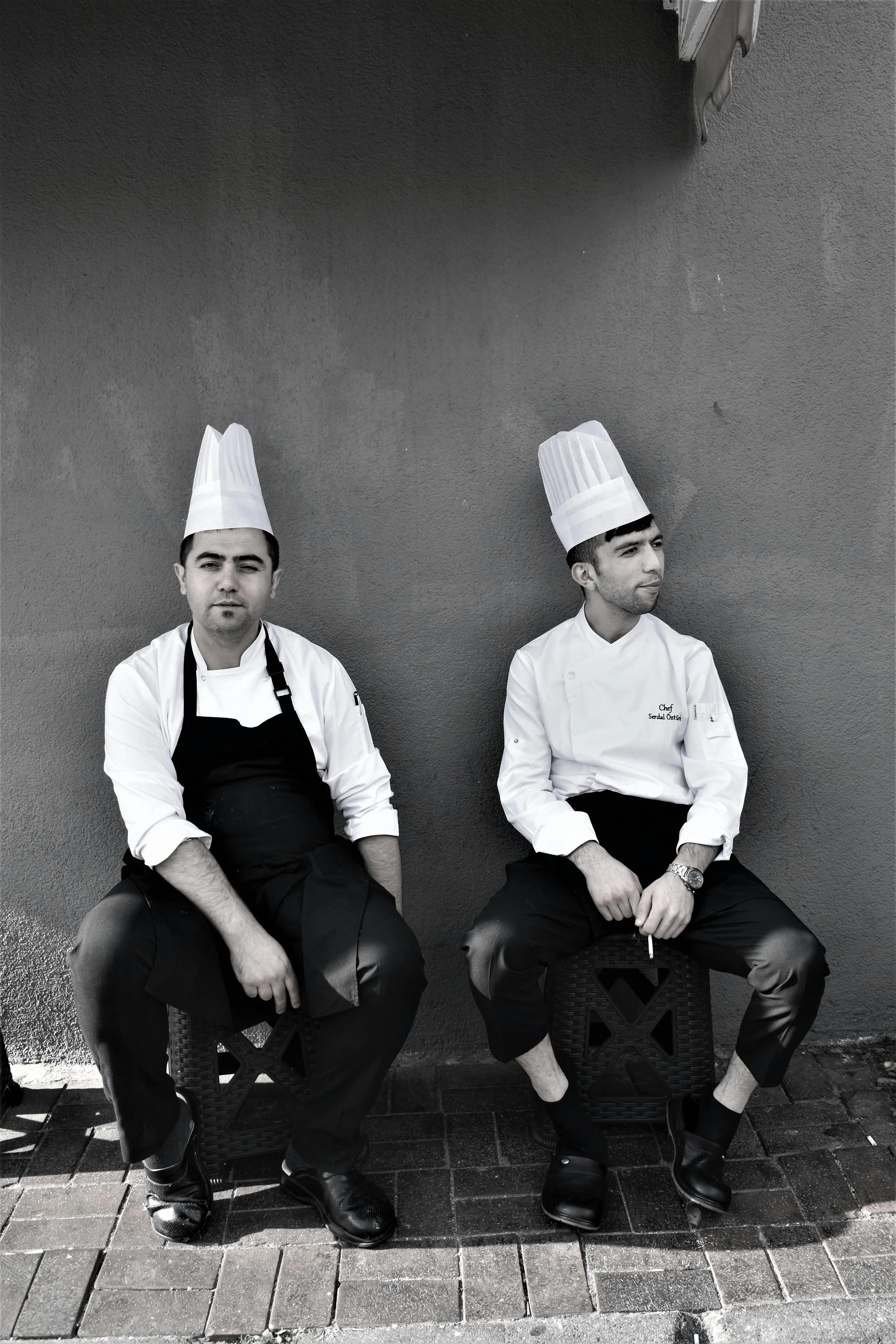 Two male chefs wearing tall white chef hats and uniforms sitting on small stools against a plain wall outdoors, one looking at the camera and the other looking away.
