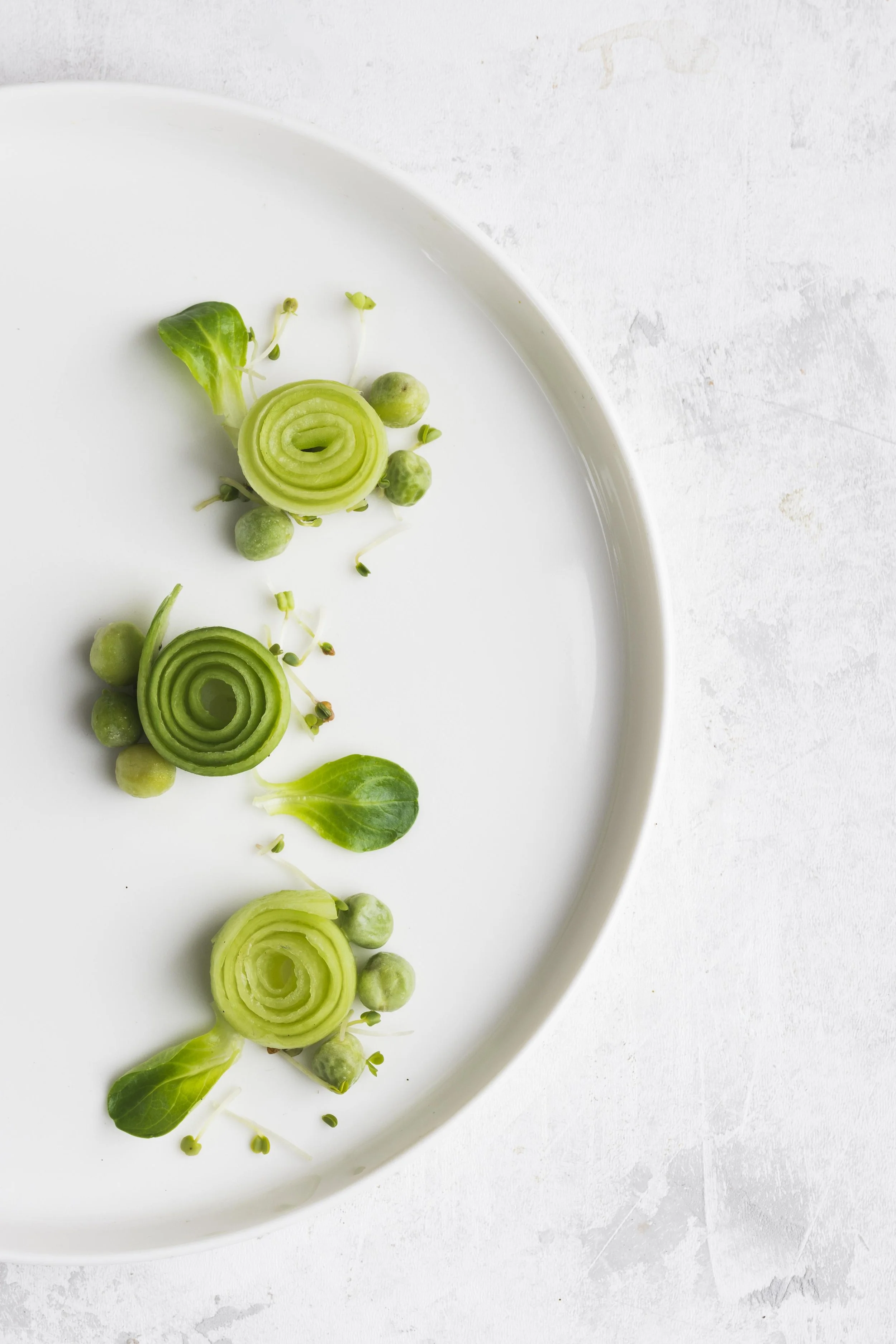 A white oval plate with sliced green vegetables arranged decoratively, including spiral slices of cucumber and small green pea pods with sprouts, on a white textured surface.