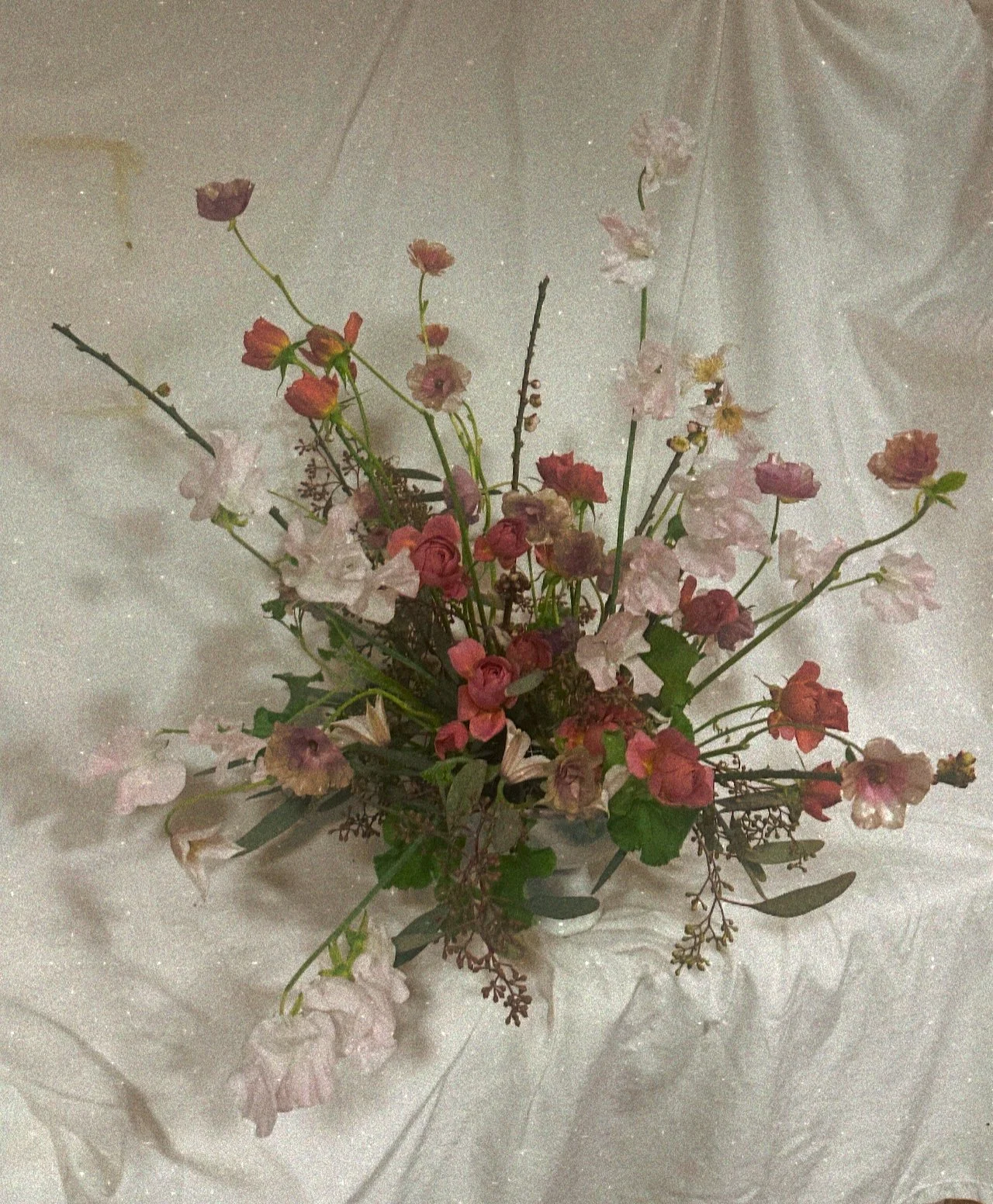 A floral arrangement with pink, red, and white flowers in a white vase, placed on a satin tablecloth with a draped fabric backdrop.