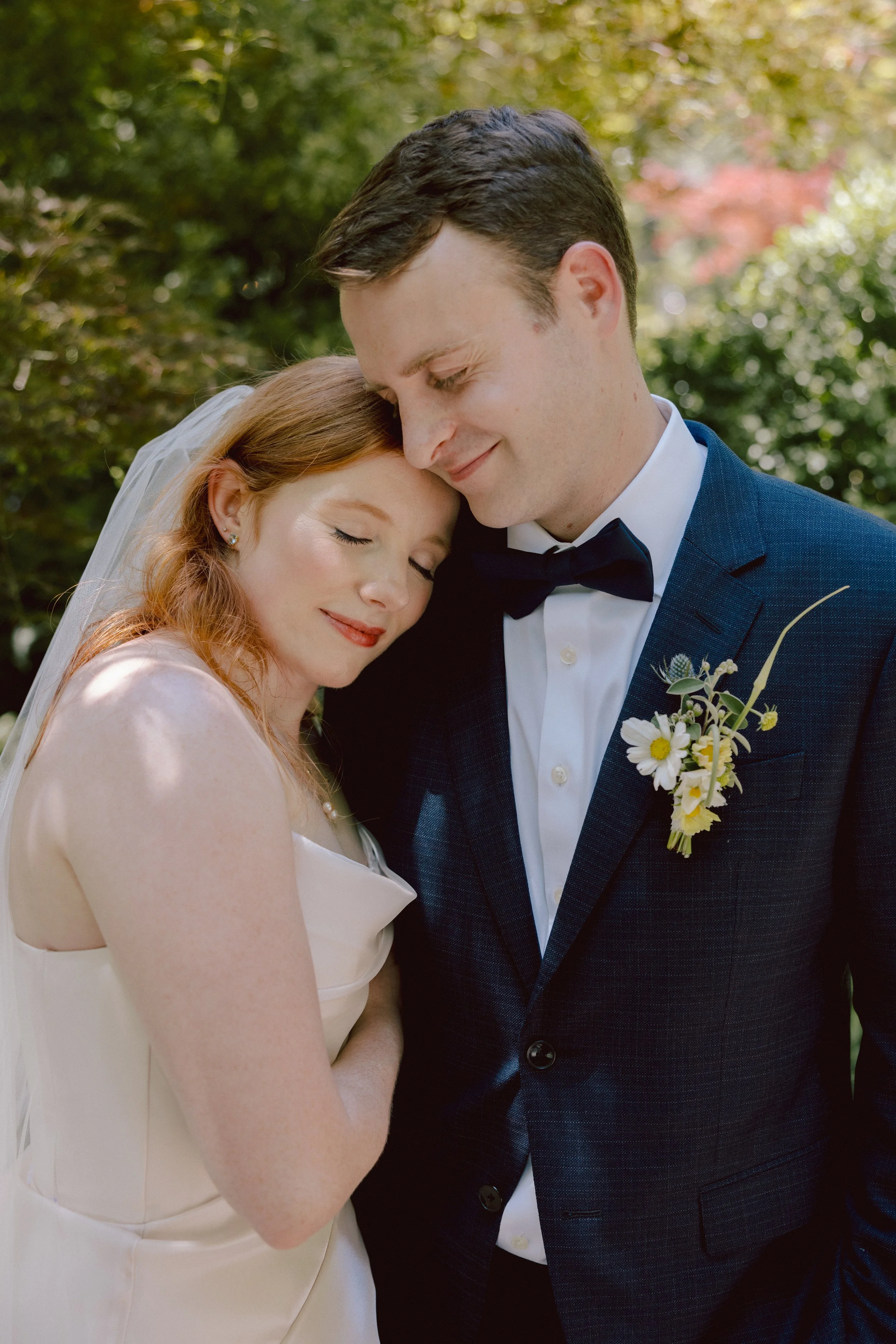 A bride and groom embracing outdoors, with eyes closed and smiling, in a garden setting with greenery and sunlight.