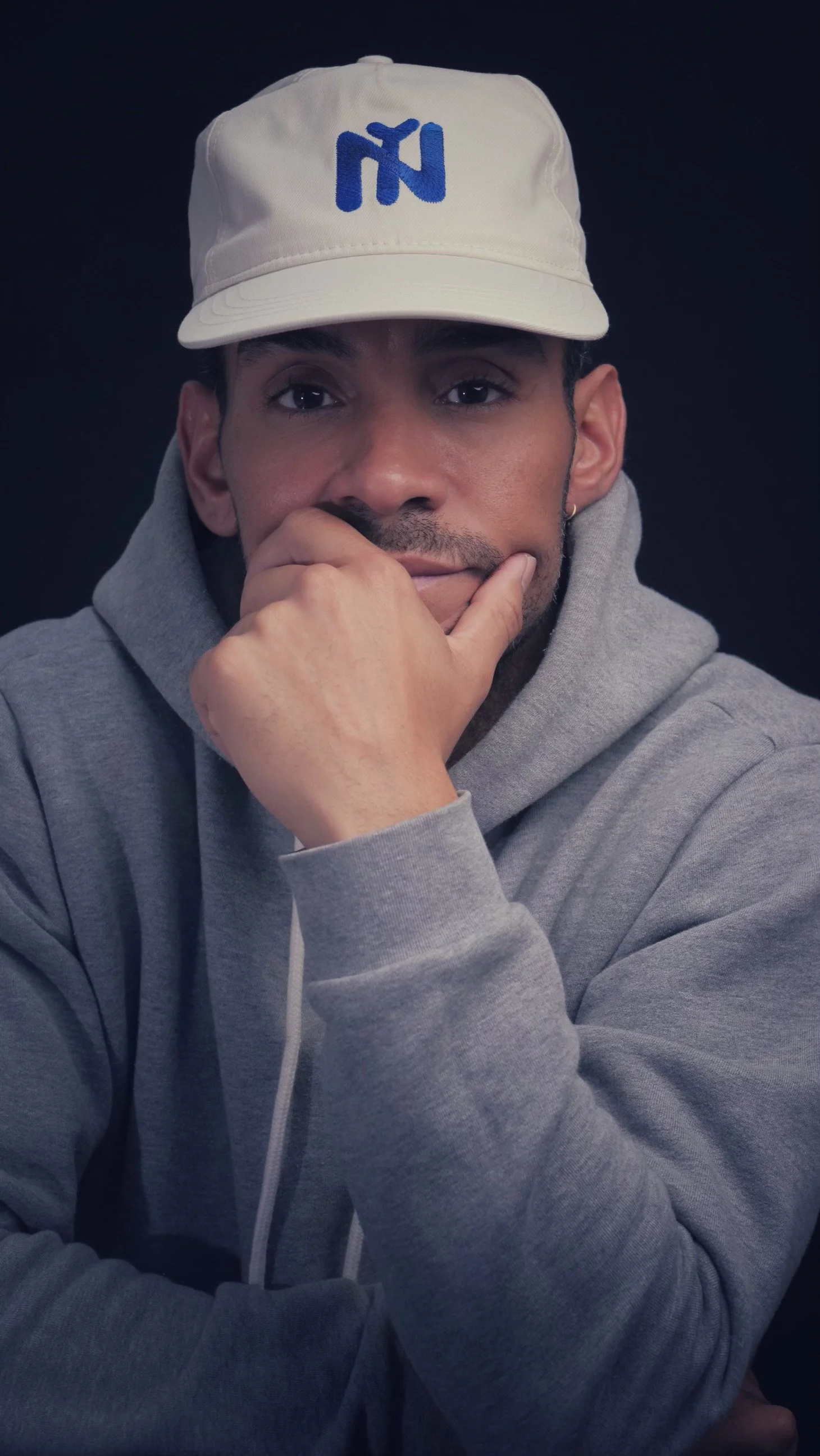 Man in gray hoodie and white cap with blue logo, resting chin on hand against a dark background.