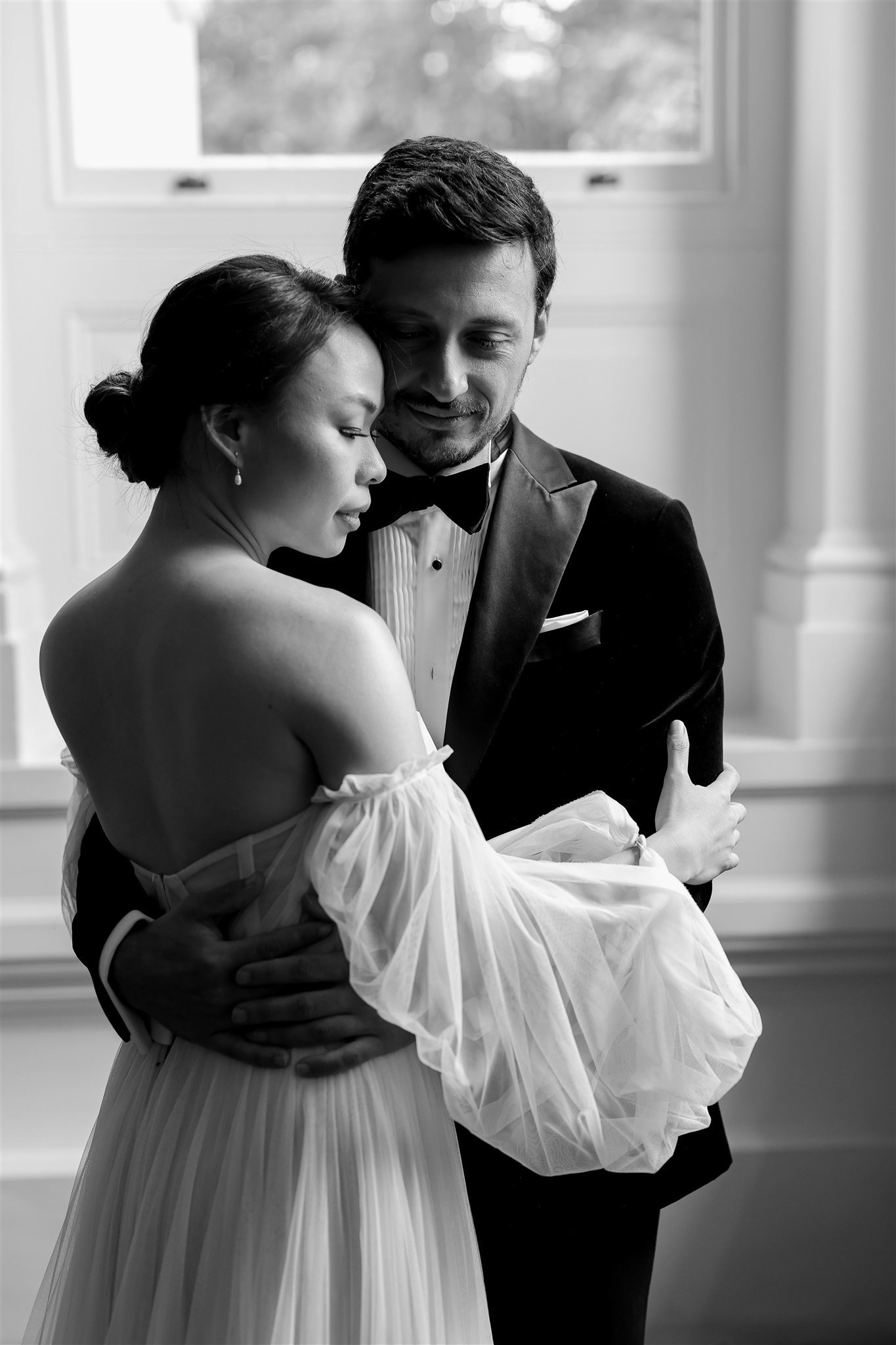 Black and white photo of an embracing couple, the woman wearing a bridal gown and the man in a tuxedo, standing in front of a window.