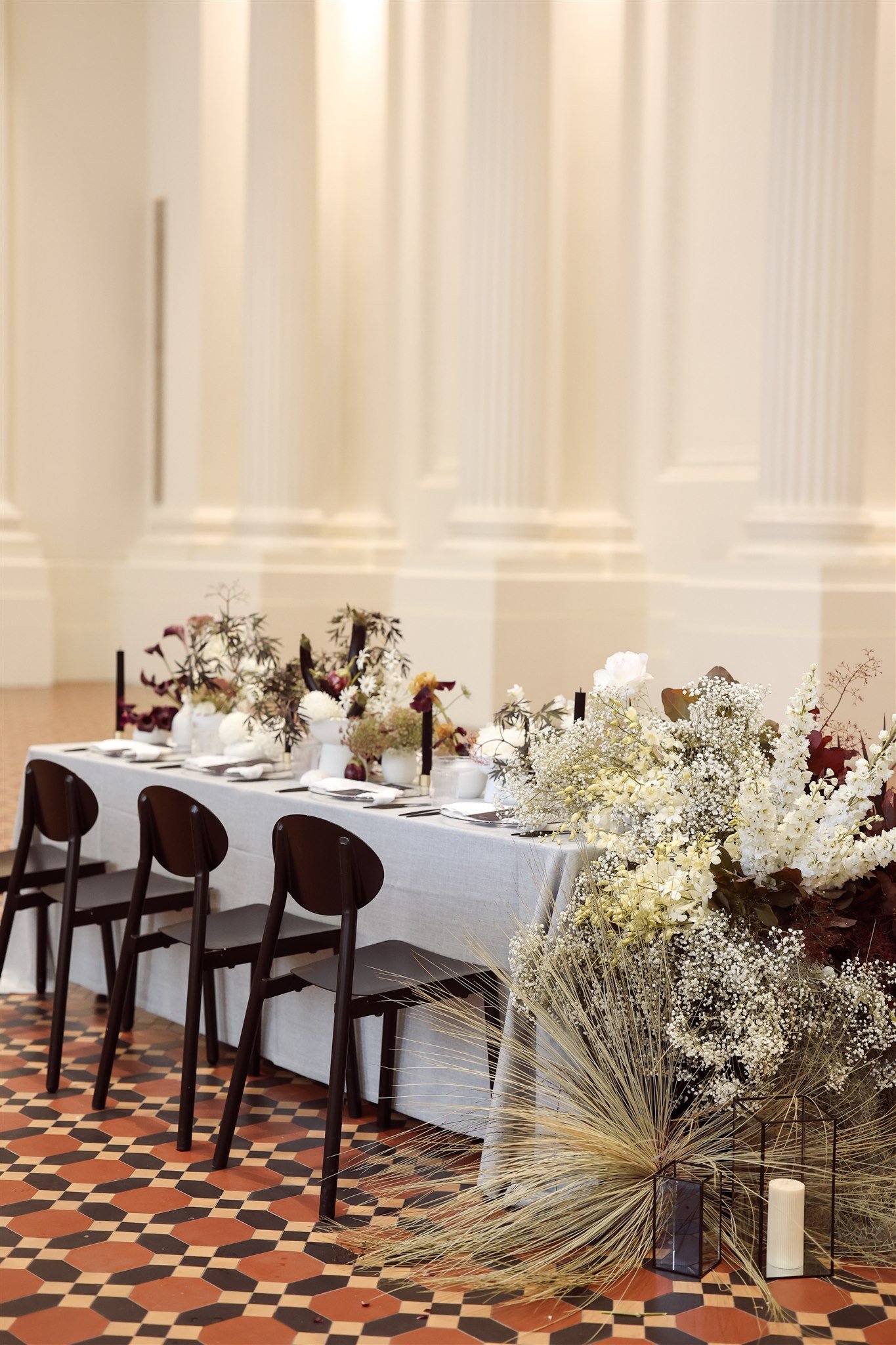 Elegant dining table setting with black chairs, floral centerpieces, and white tablecloth in a grand hall with checkered tile floor.