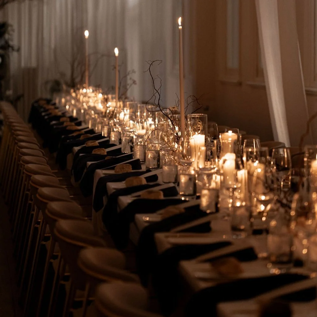 Dimly lit elegant dinner table setup with multiple rows of candles, black tablecloths, and arranged tableware.