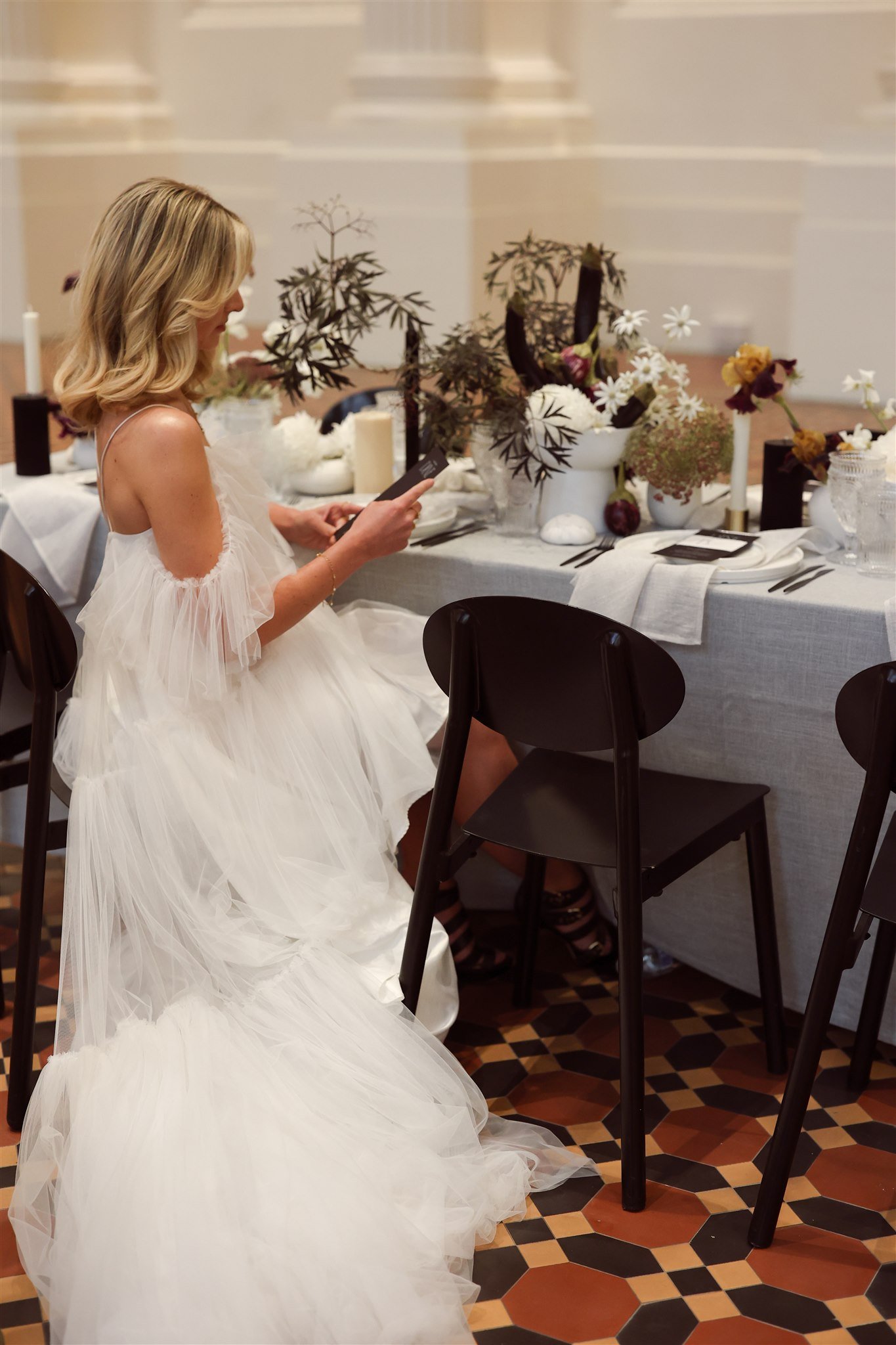 Woman in a white wedding dress sitting at a decorated table with flowers and candles, holding a card.