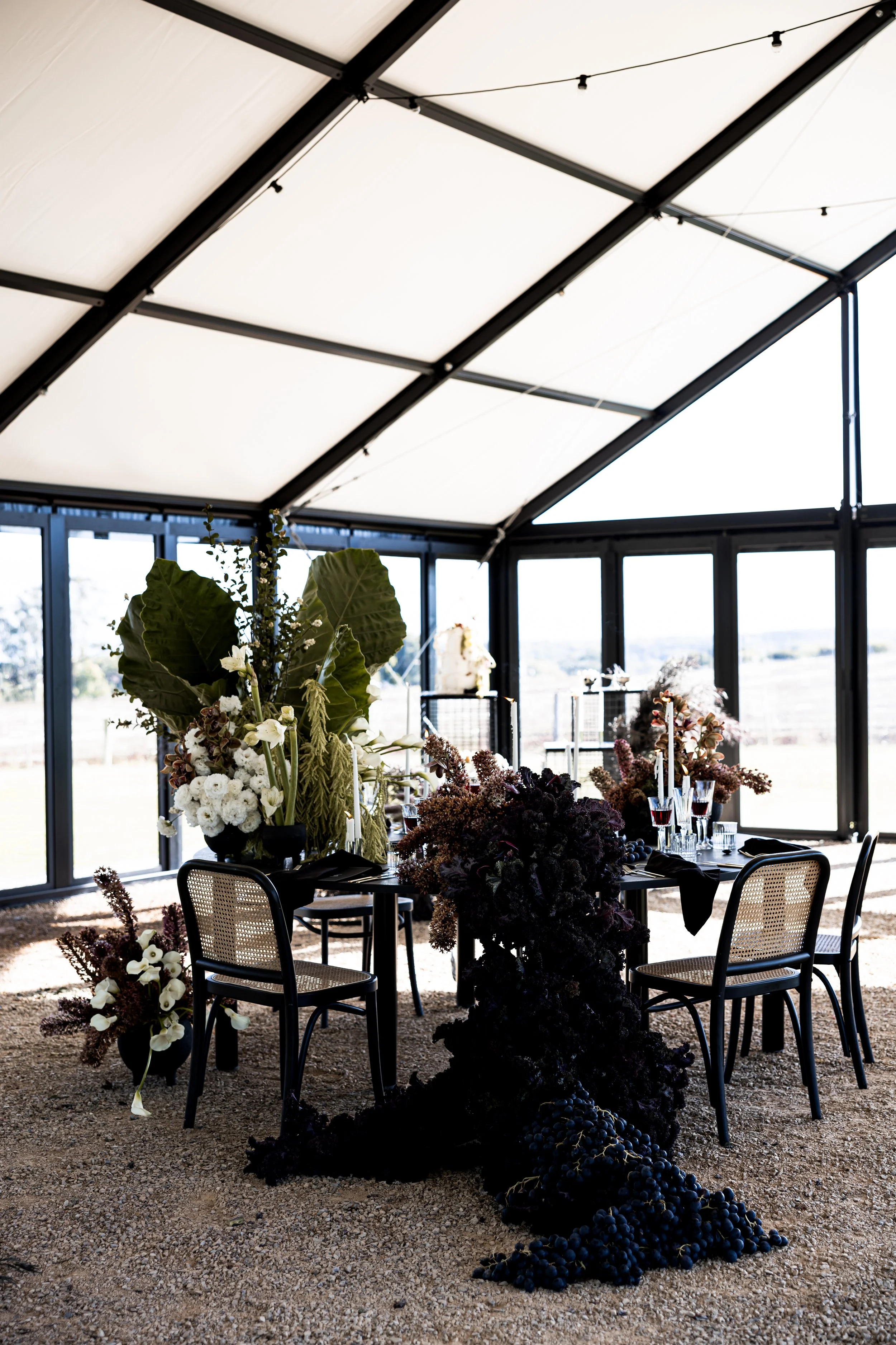 Elegant dining setup with black chairs, a table adorned with dark floral arrangements and cascading grapes, inside a glass-walled structure with a white ceiling.