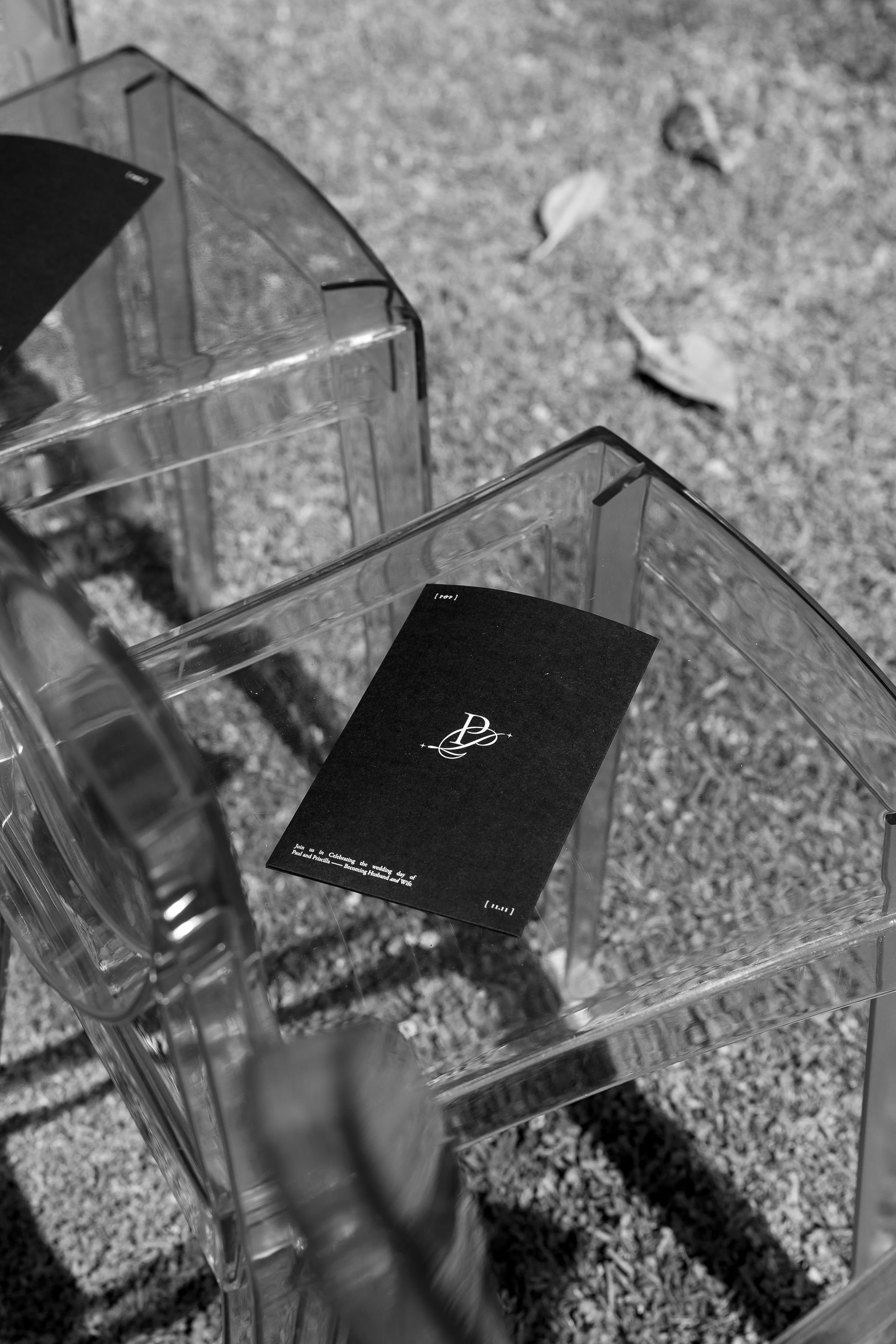 Black and white photo of transparent chairs with a black program booklet on one seat, on an outdoor surface with scattered leaves.