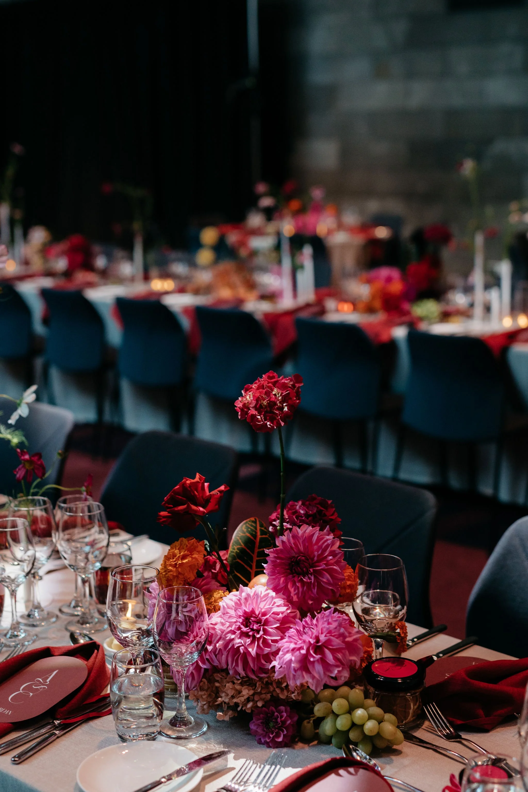 Table setting with elegant floral centerpiece featuring pink, red, and orange flowers, surrounded by wine glasses and cutlery; draped red napkins; formal dining setup.