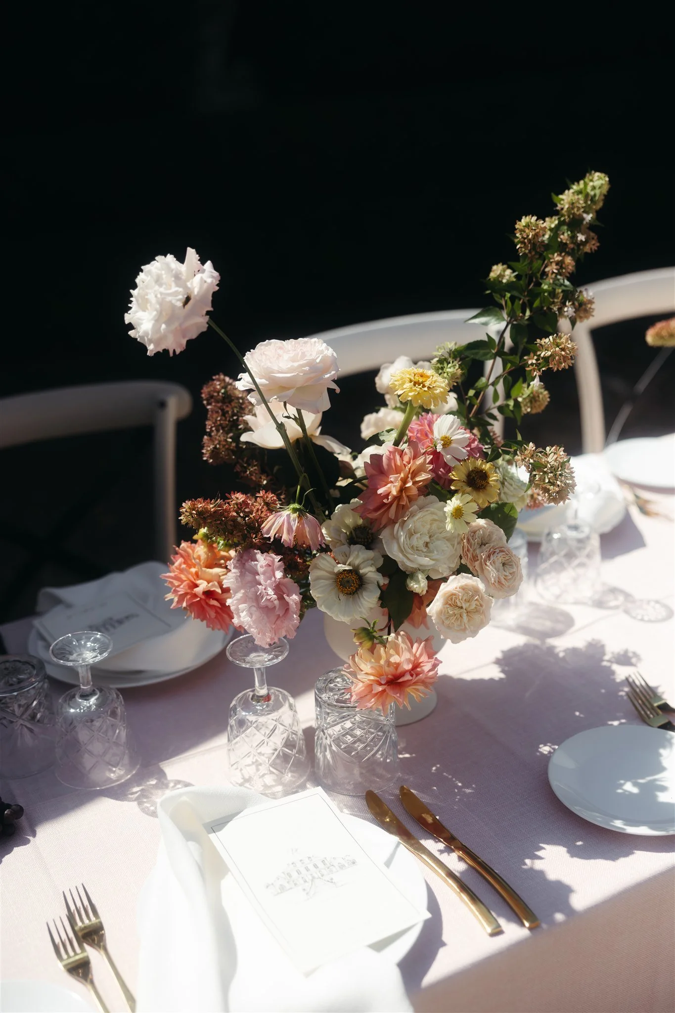 Elegant table setting with a floral centerpiece, crystal glasses, white plates, gold cutlery, and a menu on a light pink tablecloth.