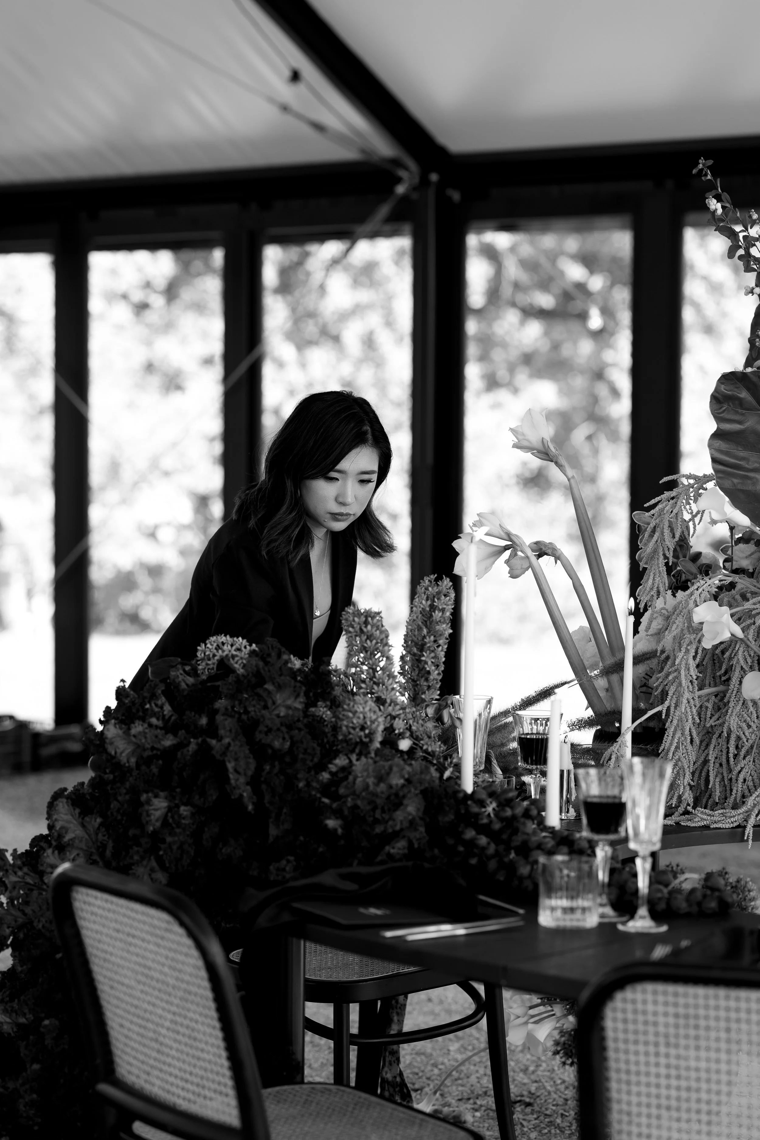 A black and white photo of a woman arranging flowers on a table set with glassware and candles in a bright indoor setting.