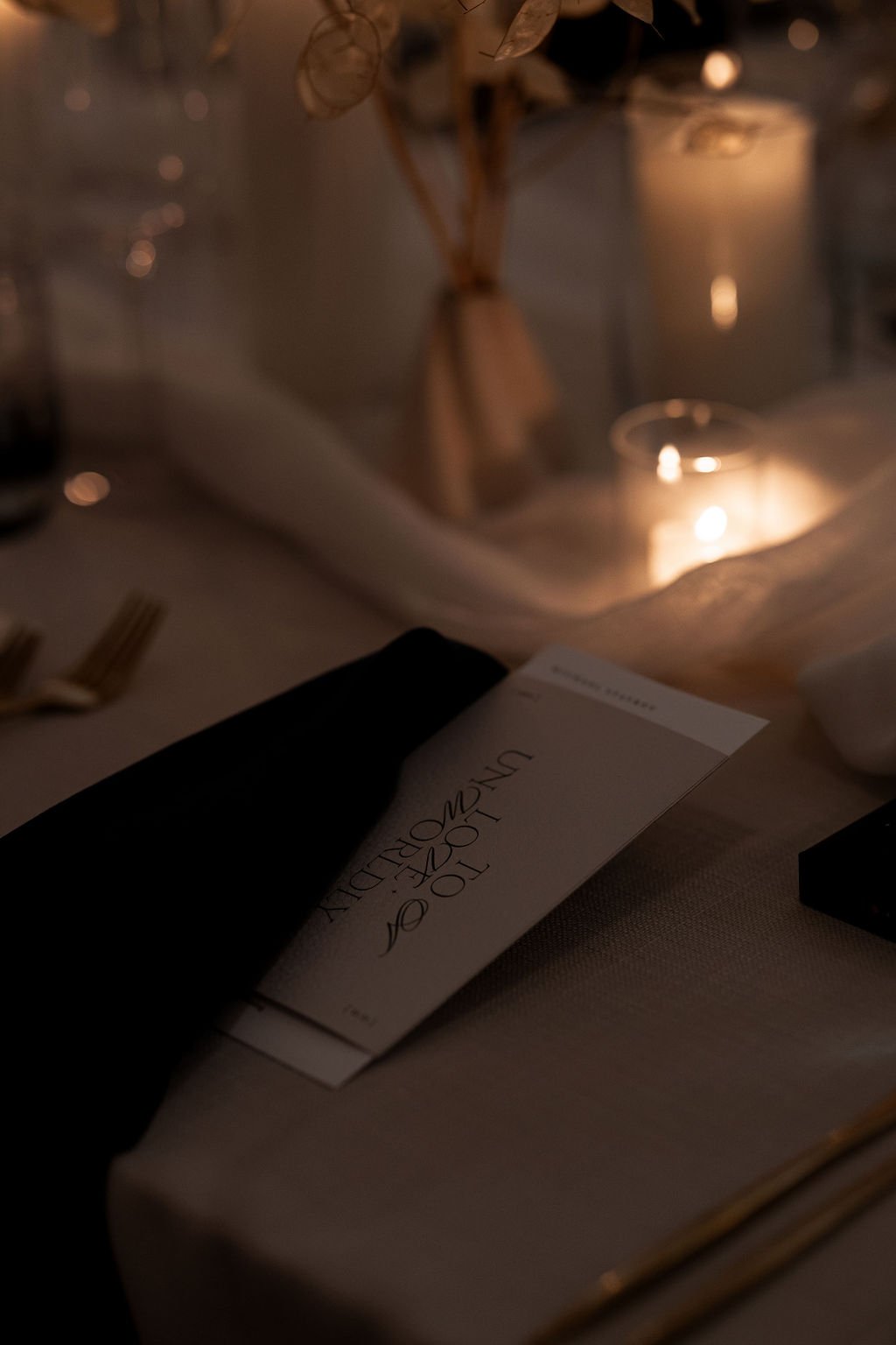 Moody table setting with a menu, black napkin, candles, and gold utensils on a beige tablecloth.
