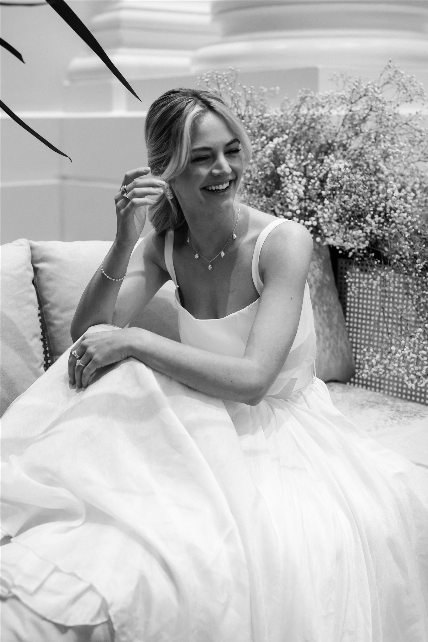 Black and white photo of a smiling woman in a white dress seated on a couch with decorative flowers in the background.
