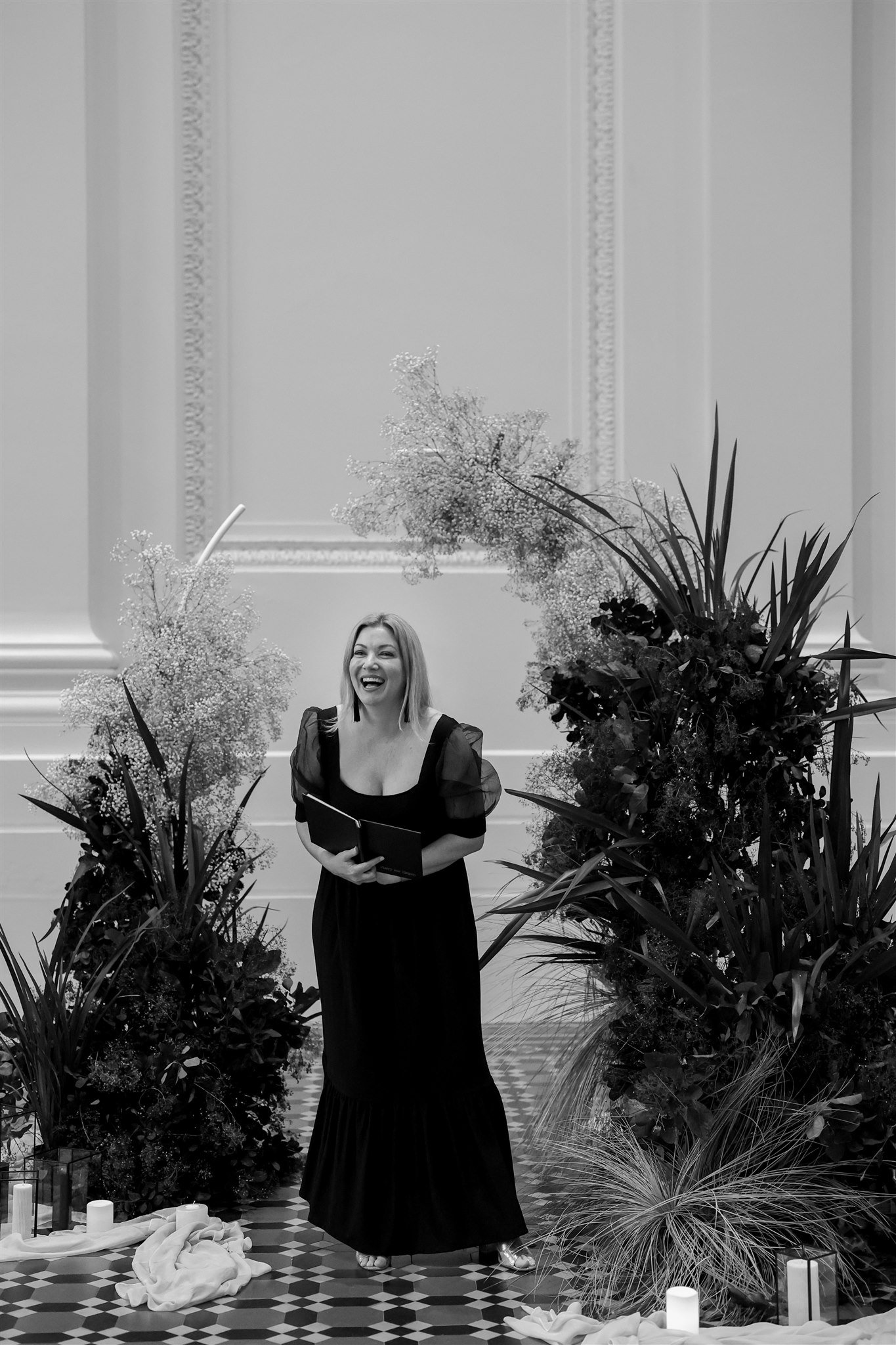 A woman in a black dress smiles, standing amidst lush greenery and floral arrangements, with candles and fabric draped on a checkered floor. The setting appears indoors with ornate wall details.
