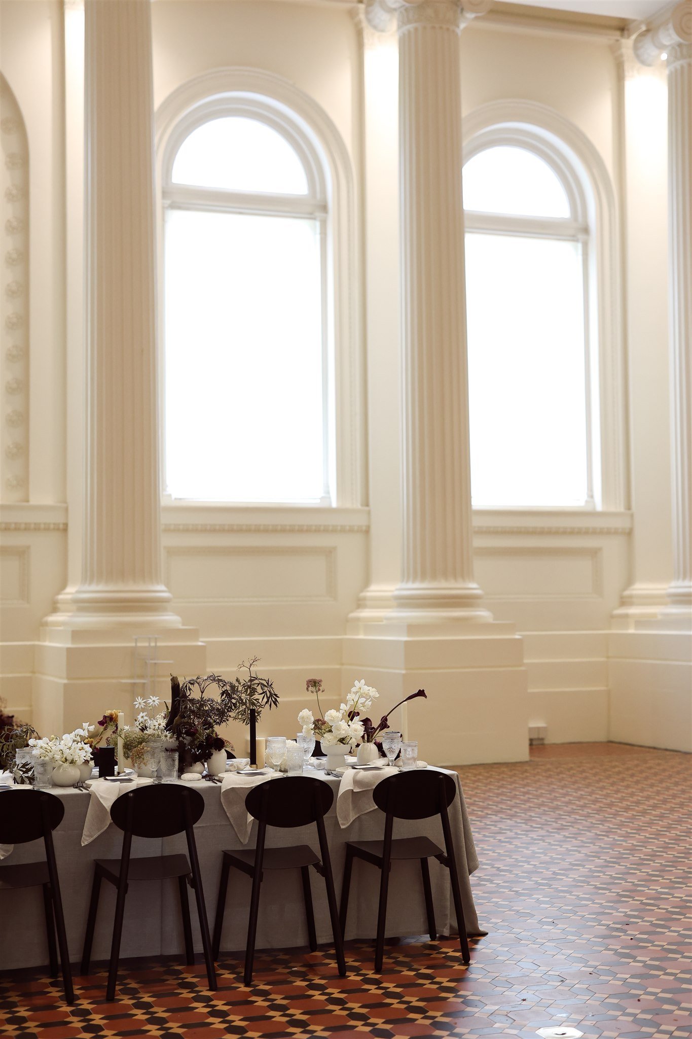 Elegant dining table setup in a grand hall with tall windows and columns, featuring floral centerpieces and tableware.