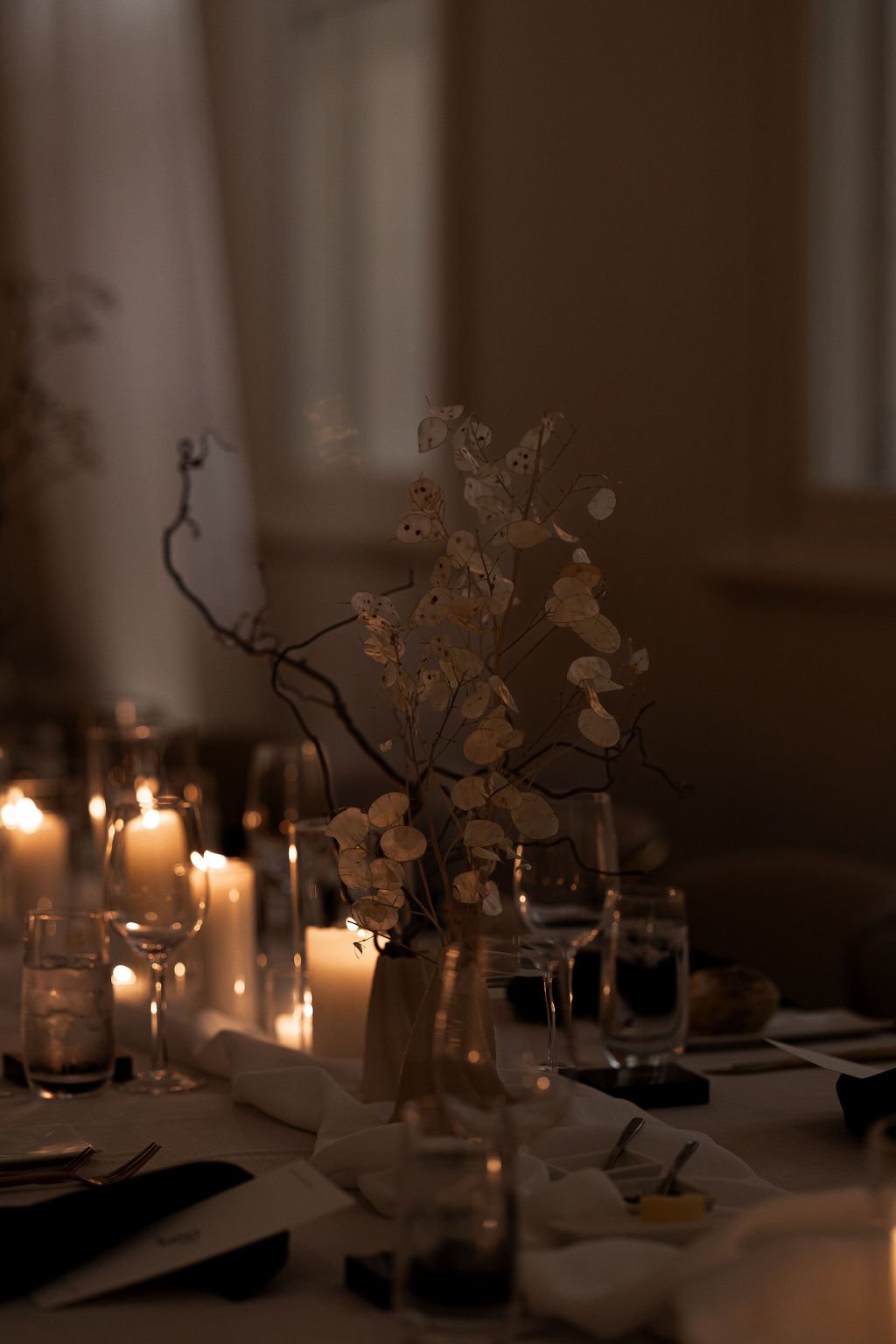 Dimly lit dining table with candles, glassware, and a centerpiece of dried branches in a vase.