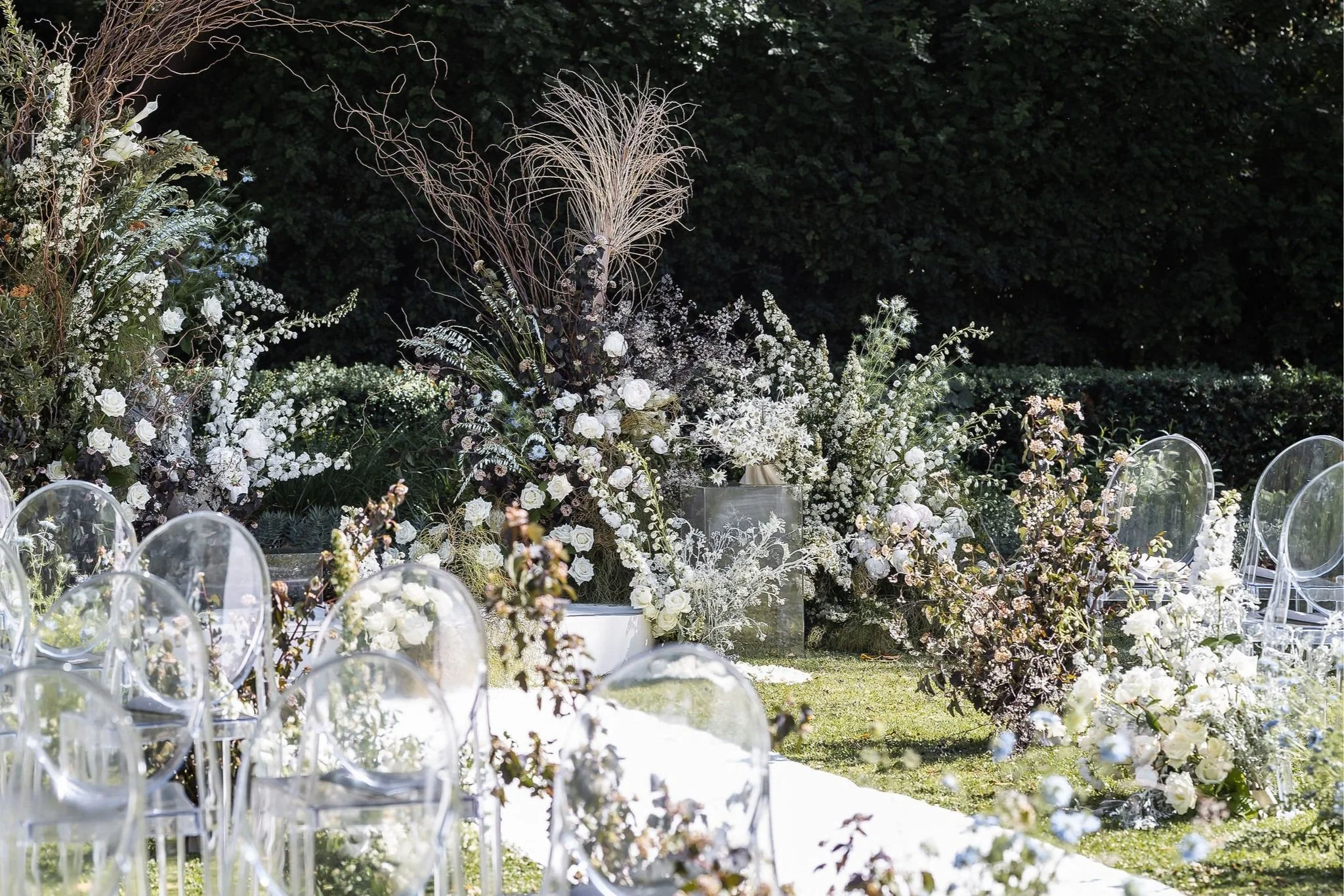 Outdoor wedding ceremony setup with clear chairs and floral decorations, featuring white and green flowers, against a lush garden backdrop.