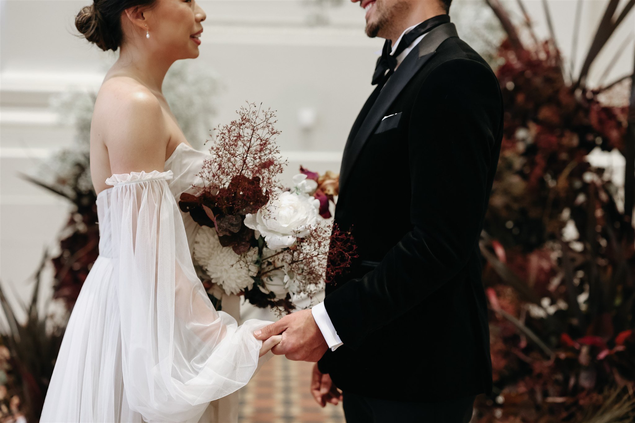Bride in a white dress holding a bouquet stands facing a groom in a black tuxedo, holding hands, with blurred floral arrangements in the background.