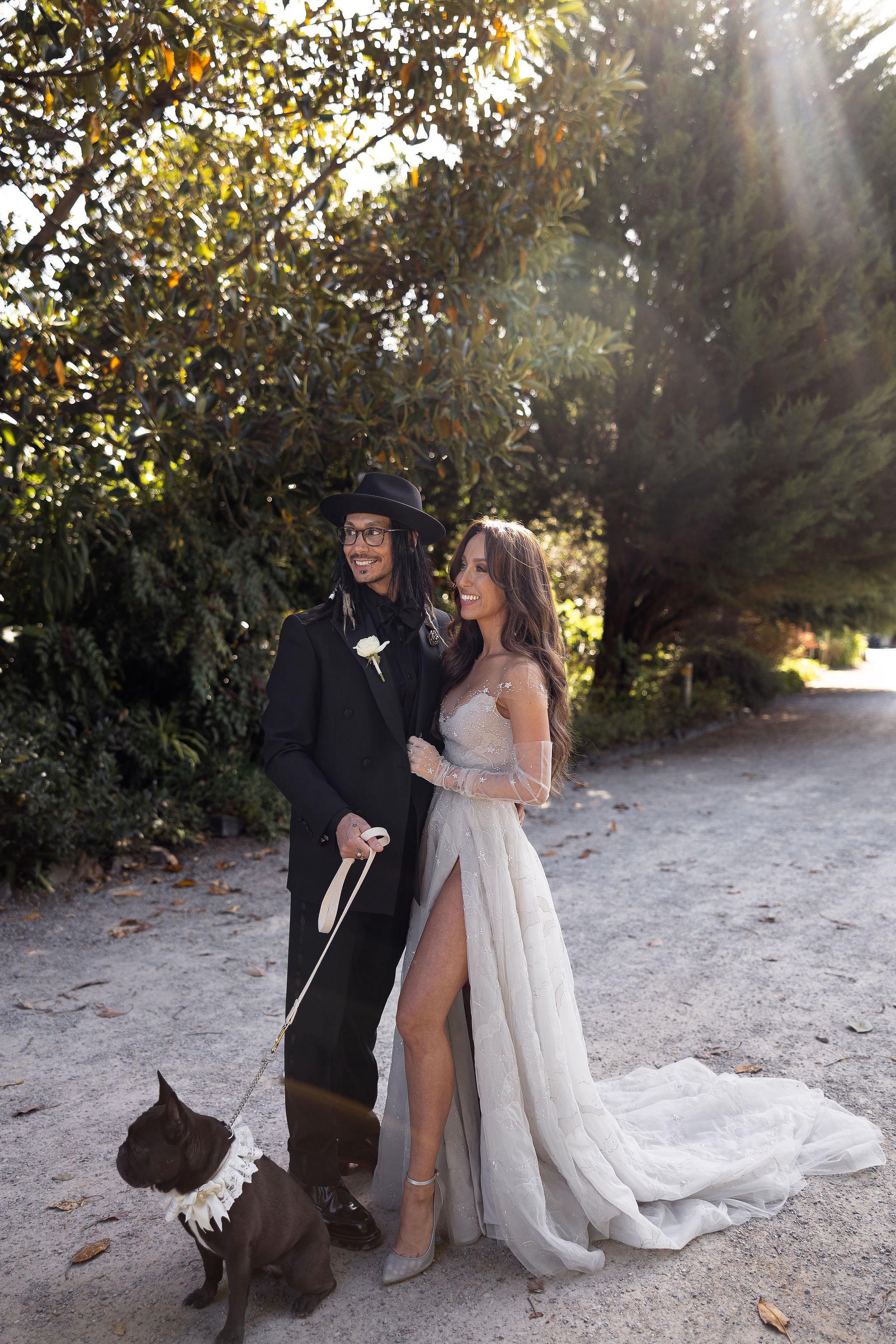 A couple in formal wedding attire with a French bulldog, standing outdoors in a garden.