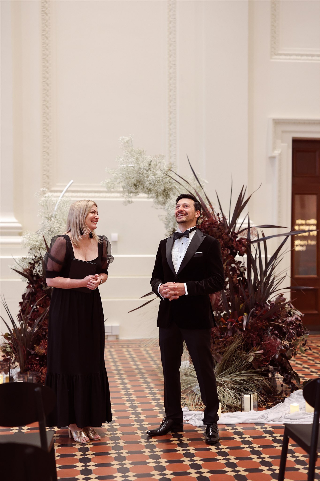 A man and woman dressed in formal attire stand smiling in a room with elegant decor and floral arrangements.