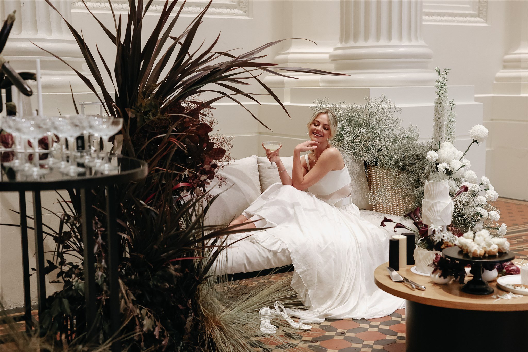 A woman in a white dress reclining on a couch, holding a drink, surrounded by plants and flowers, with a table of desserts in the foreground and columns in the background.
