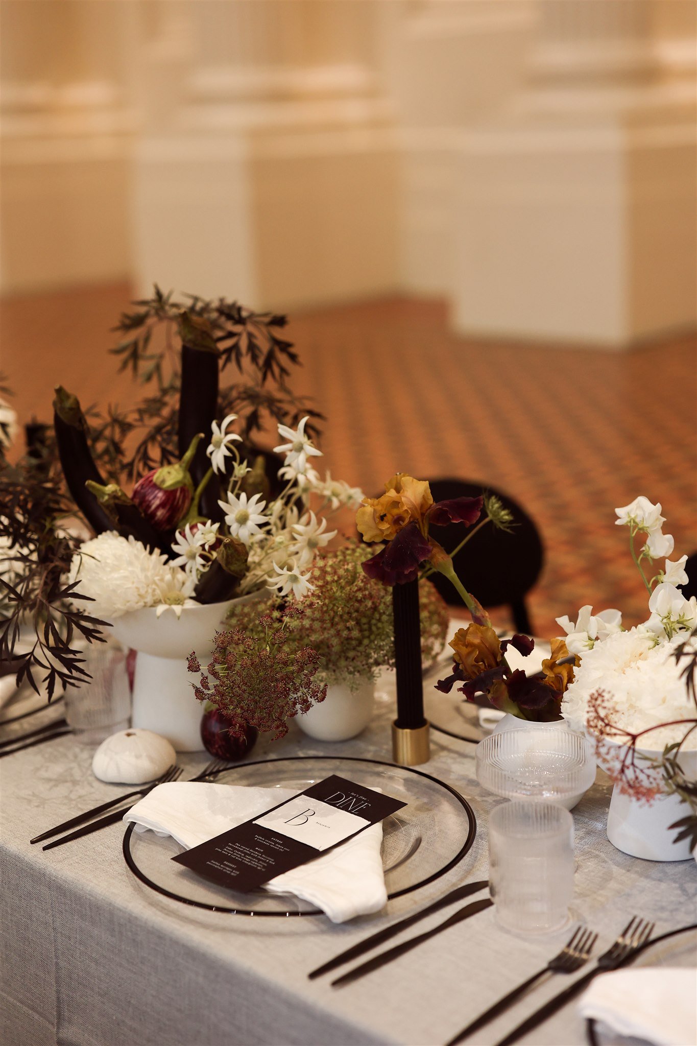 Elegant table setting with floral centerpiece, black candle holders, and clear glassware on a gray tablecloth, featuring a menu card placed on a plate.