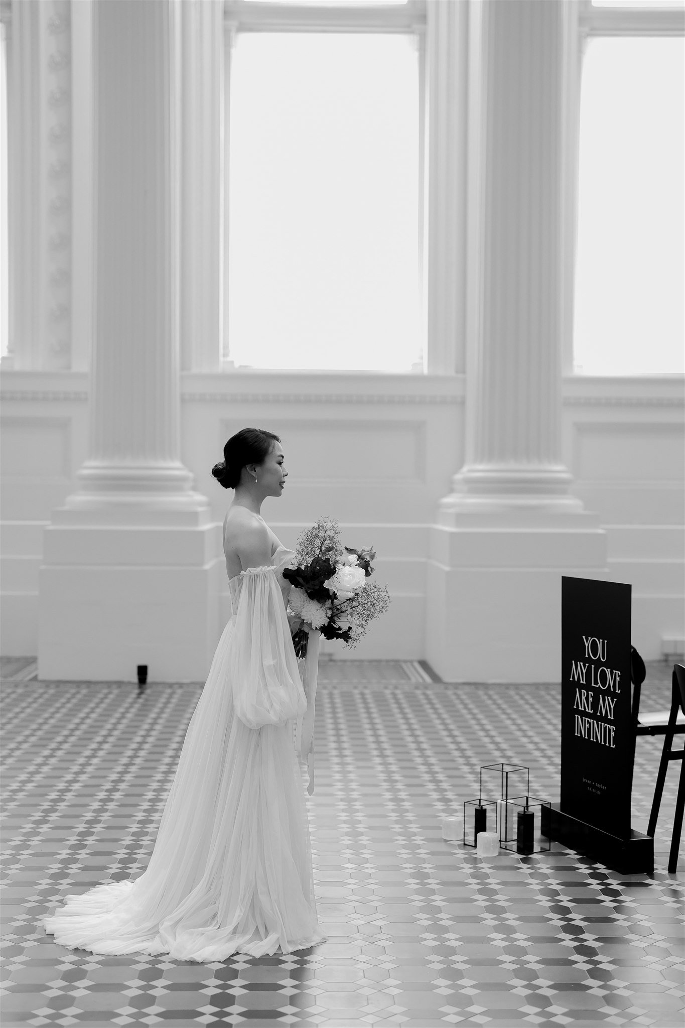Bride in a flowing gown holding flowers in a grand, white interior. Large columns and checkered floor are visible, with a sign reading "You my love are my infinite."