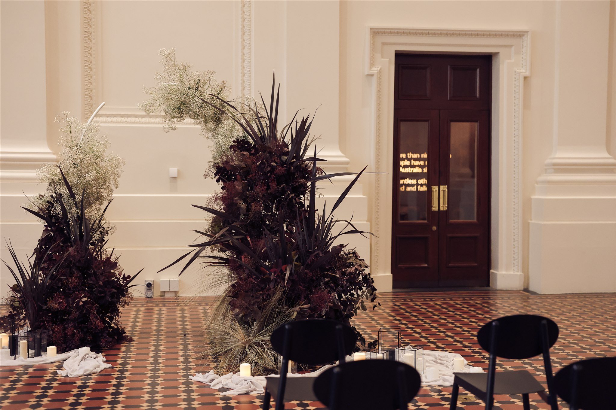 Interior with floral arrangements, black chairs, candles, and decorative tiles in a large room with wooden double doors.