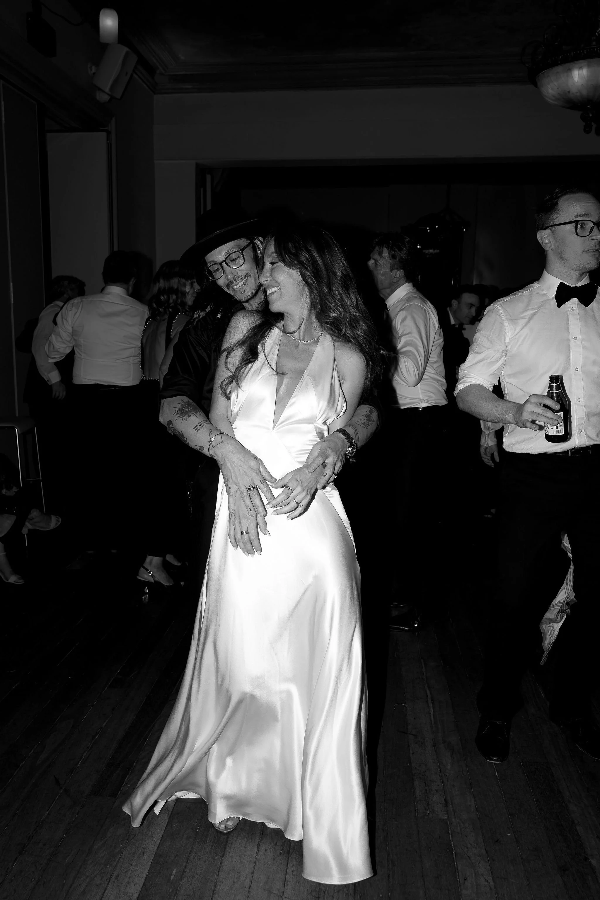 A black and white photo of a couple dancing closely at a party. The woman is wearing a satin gown and the man is embracing her from behind. Other partygoers are visible in the background. One man is holding a beer bottle.