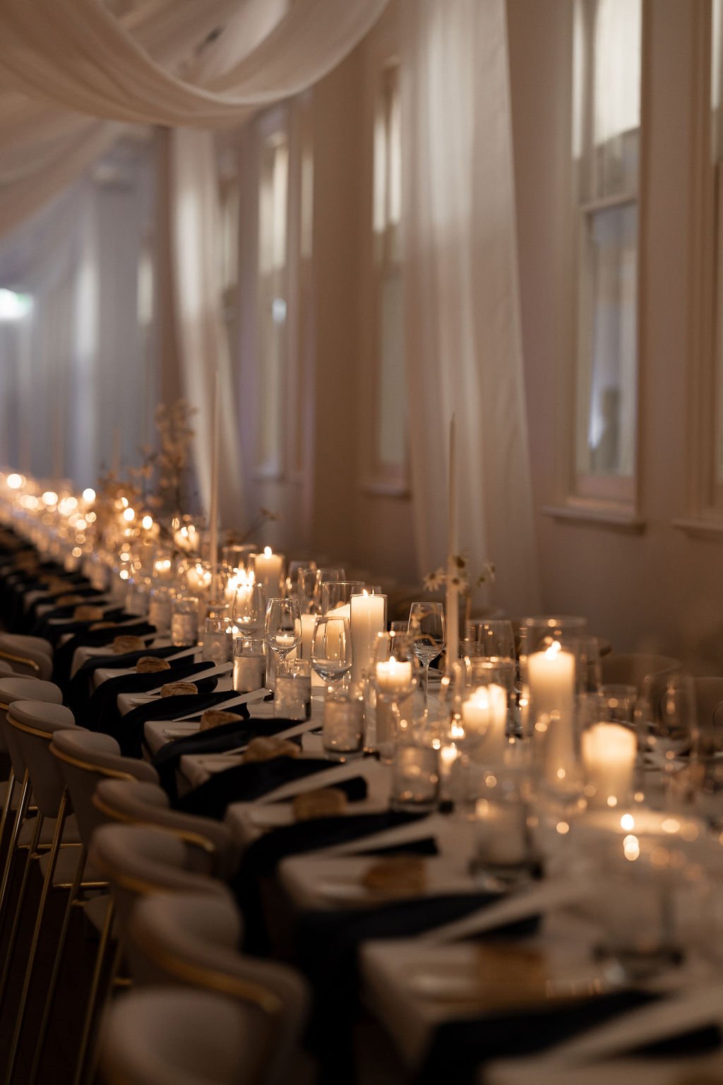 Elegant table setting with candles and glassware, featuring black napkins on white tablecloths. Ambient lighting creates a warm atmosphere, with draped fabric overhead.