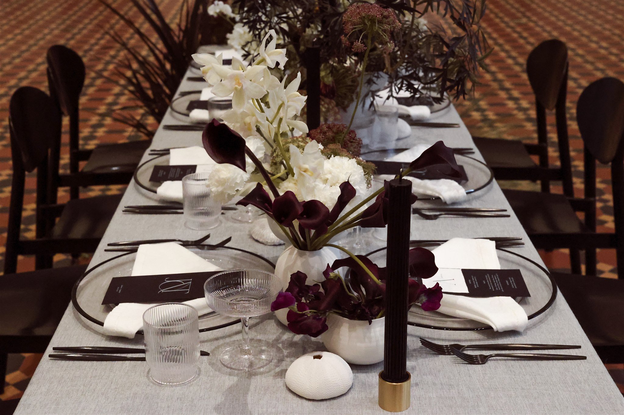 Elegant dining table set with dark flowers, white tablecloth, clear glass dinnerware, black cutlery, and decorative vases.