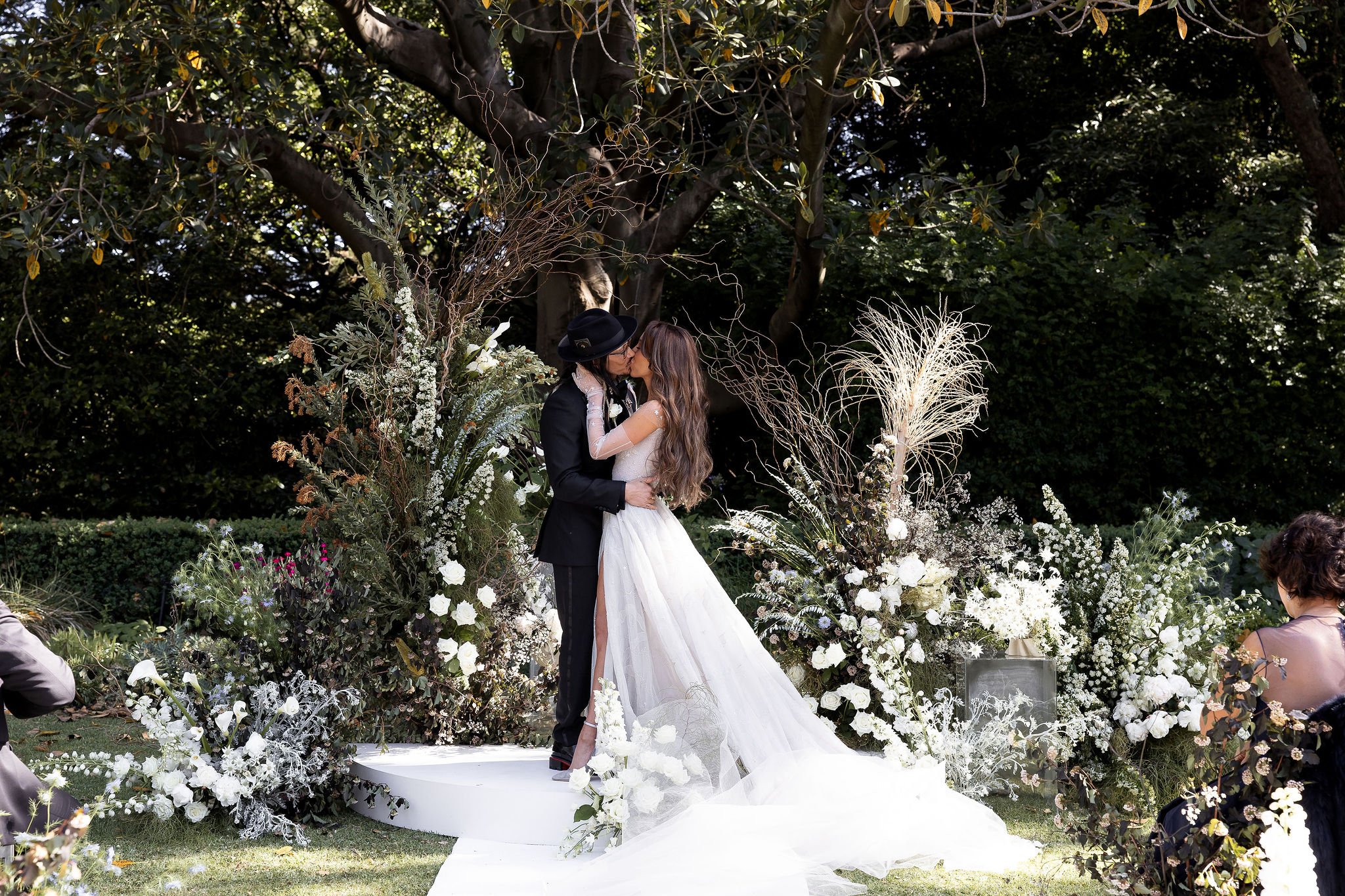 Bride and groom kissing at outdoor wedding ceremony, surrounded by white flowers and greenery, under a large tree.