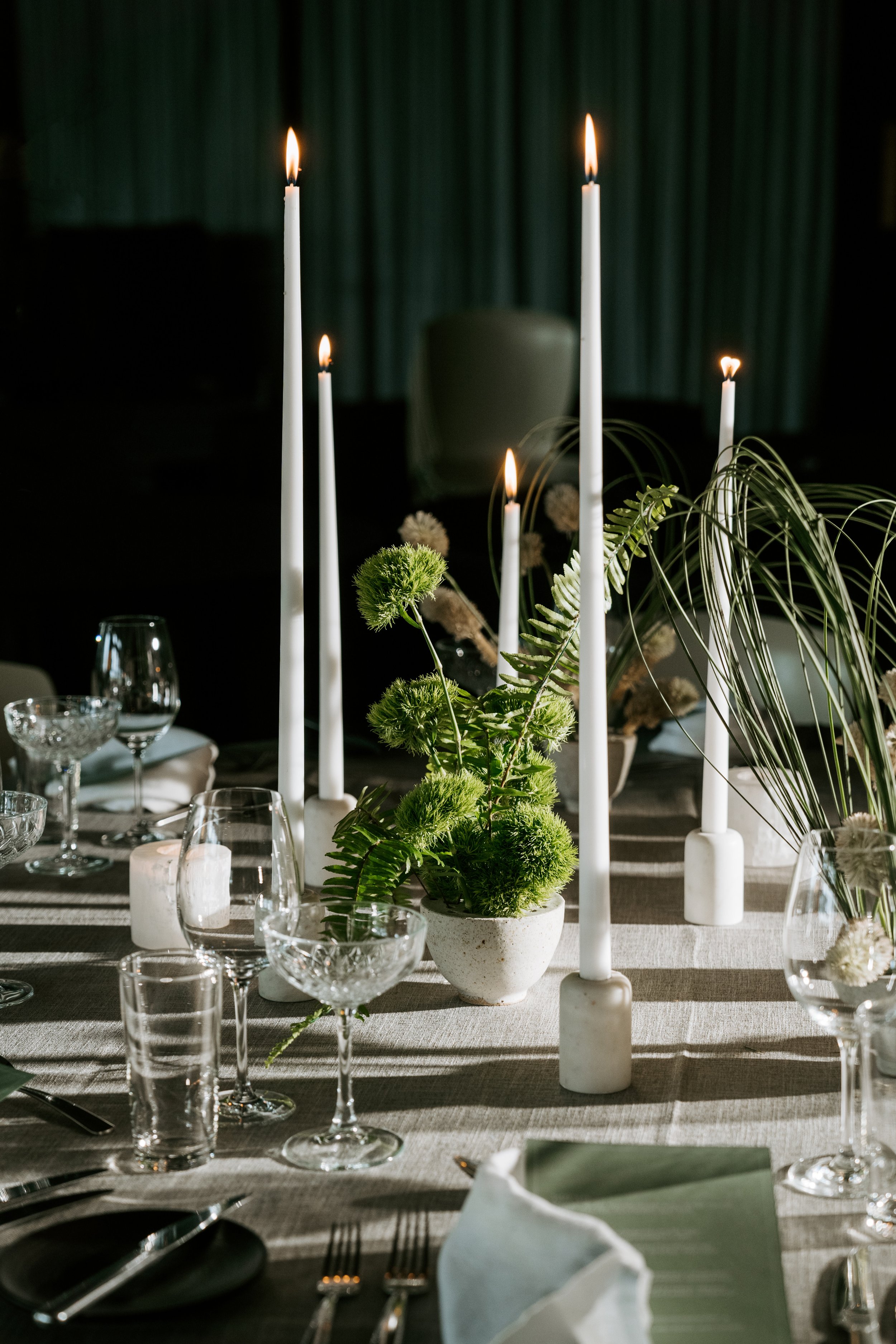 Elegant table setting with lit taper candles, green floral centerpiece, crystal glassware, and white napkins on a linen tablecloth.